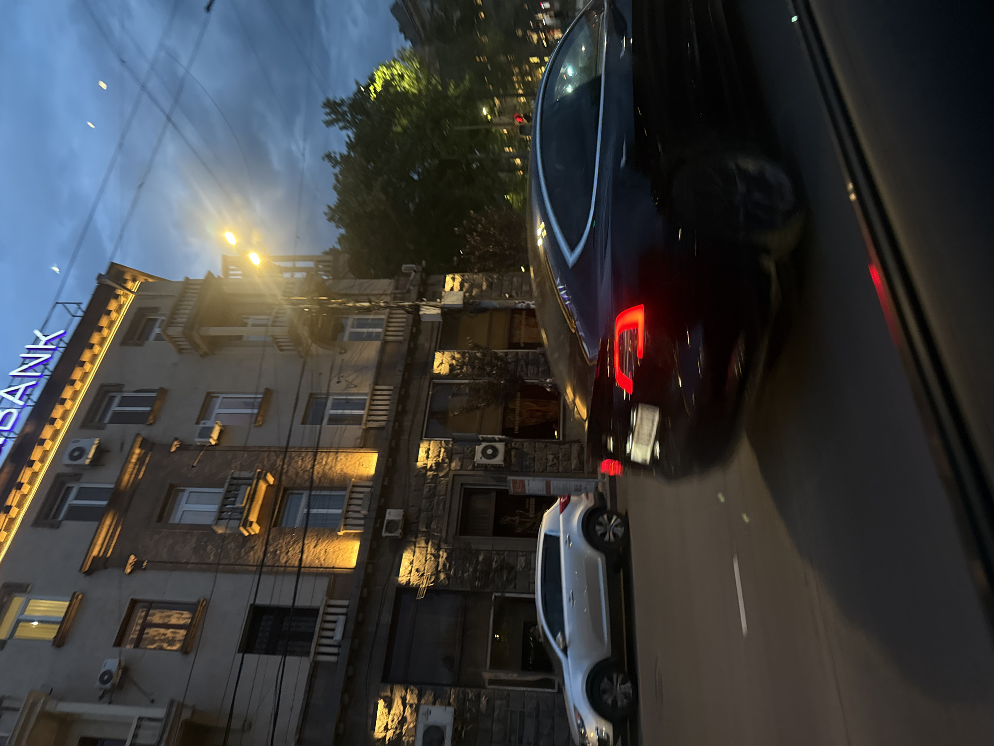 The image shows a nighttime scene in Yerevan, Armenia. The view is taken from inside a vehicle, likely a car, looking out onto a street.
On the left, there is a multi-story building with a sign that reads "Bank" in Armenian. The building has numerous windows, some of which have visible air conditioning units. The facade of the building appears to be a mix of stone and plaster.
On the street, there are two vehicles: a dark-colored car immediately to the right of the frame, partially obscuring the view, and a light-colored car further down the road. Streetlights are visible, illuminating the buildings and the road.
The sky is dark, suggesting that it is either late evening or night. Some trees are visible in the background between the buildings.