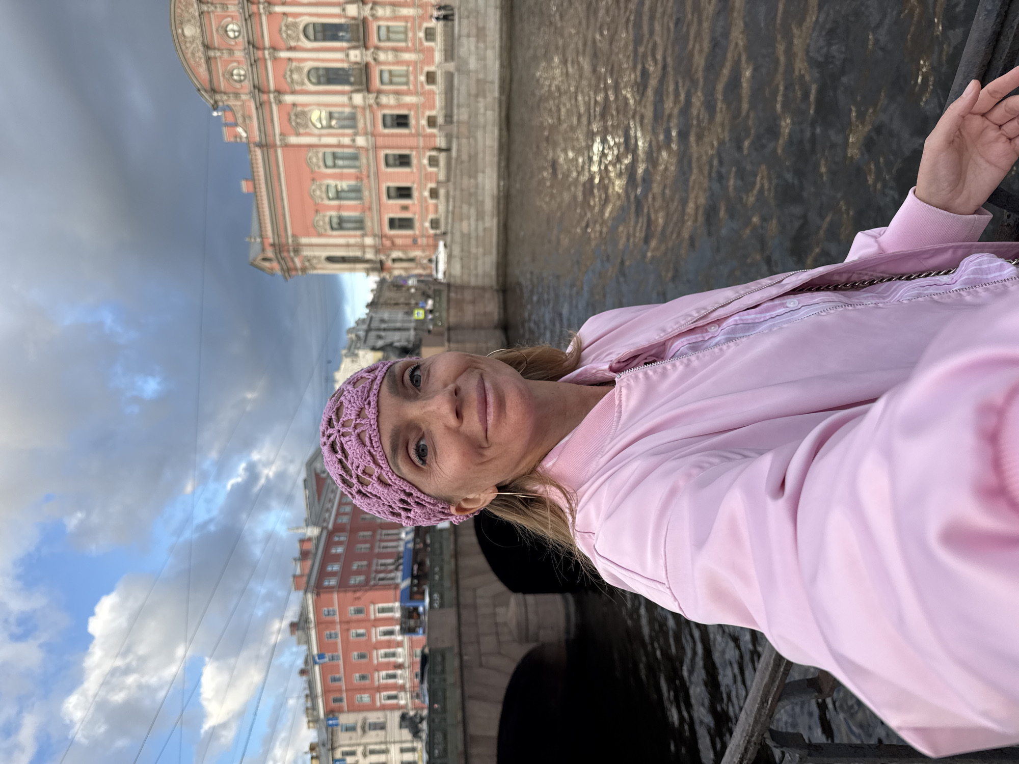 In a selfie taken outdoors, a fair-skinned woman with blonde hair pulled back and wearing a pink crocheted hat and a pink bomber jacket smiles at the camera. She is positioned in the foreground, with a river and city buildings in the background. The buildings appear to be in a European style, with ornate architecture. The sky is cloudy with patches of blue. The overall scene suggests a candid moment captured during a trip. The location is likely Saint Petersburg, Russia, given the architectural style and the presence of a canal or river within a city.