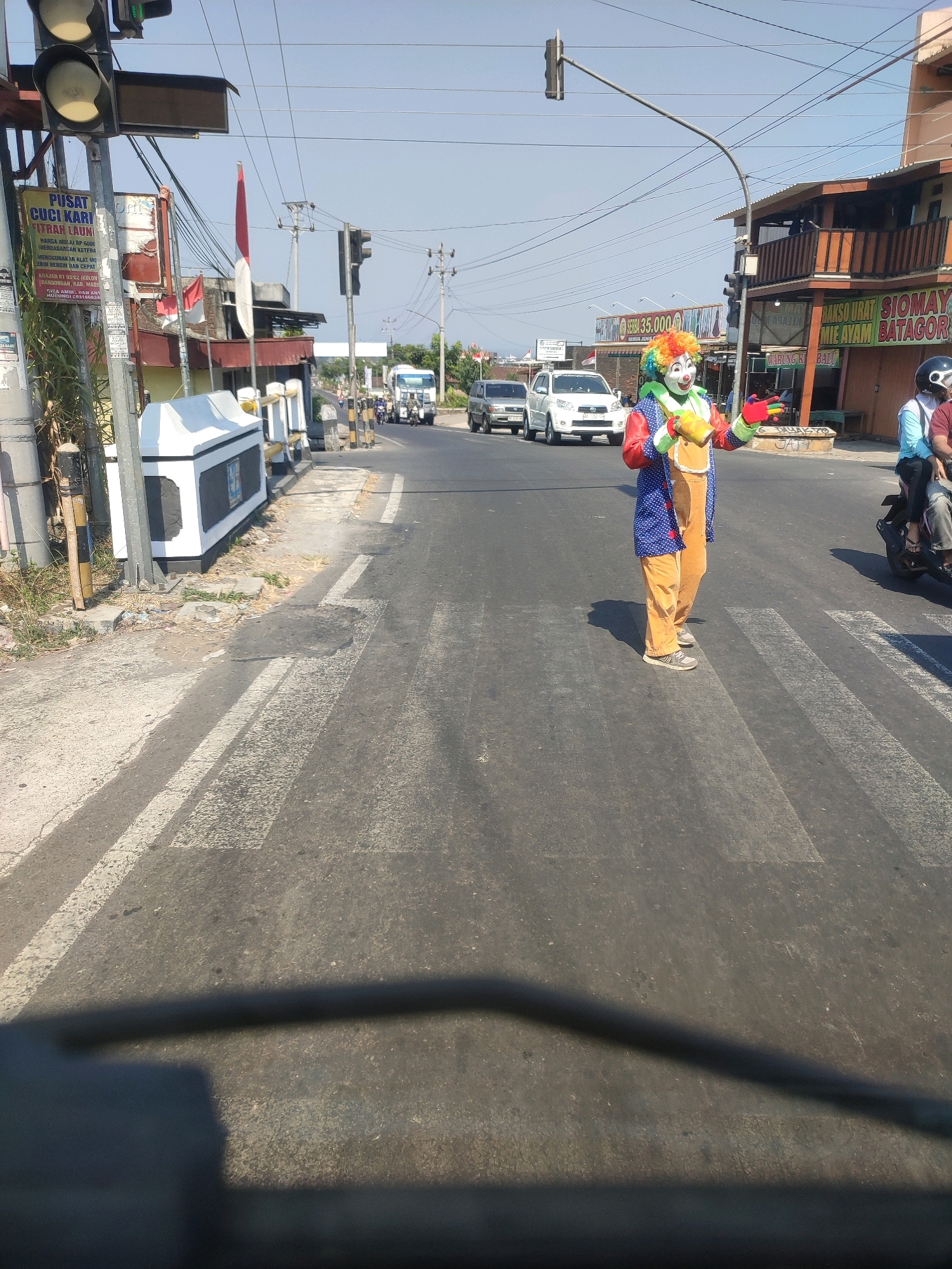 The image depicts a street scene in Trasan, Indonesia, on a sunny day. In the foreground, a person dressed as a clown stands on a pedestrian crossing. The clown is wearing a colorful wig, face paint, a blue jacket with white polka dots, orange overalls, and grey shoes. They are holding a yellow object and appear to be interacting with the traffic. Behind the clown, several cars and motorcycles are visible on the road. A building with signs in Indonesian is located on the right side of the image. Traffic lights and utility poles are also present in the background. The scene is set on a paved road with white pedestrian crossing lines.