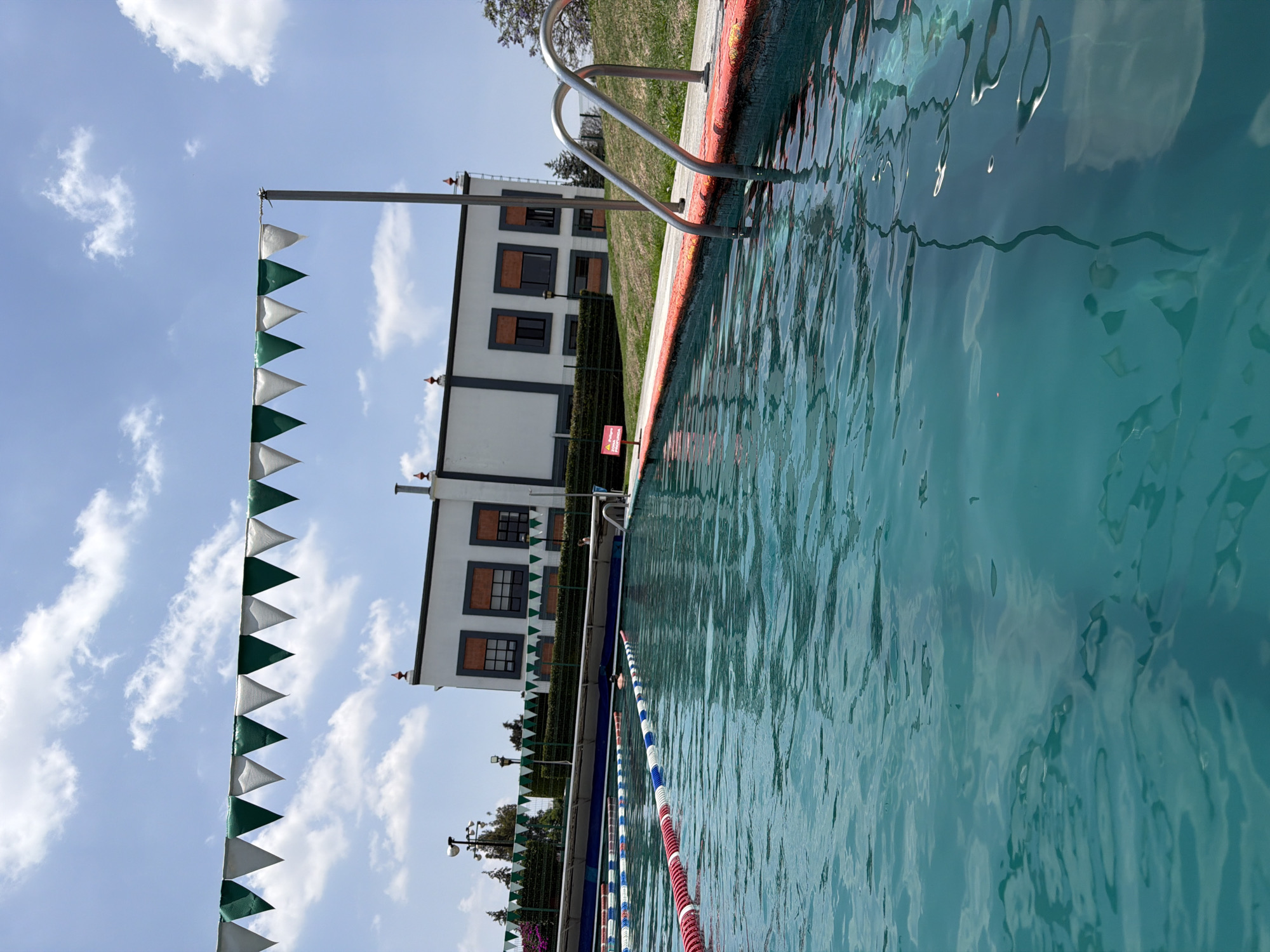 The scene shows a swimming pool with a metal ladder leading into the water. The pool's edge is marked with red and white paint. Dividing ropes, striped blue, white, and red, float in the water. A white building with dark-framed windows stands next to the pool, adorned with green and white triangular pennants. The sky is blue with scattered clouds. The scene is located in San Andrés Cholula, Mexico.
