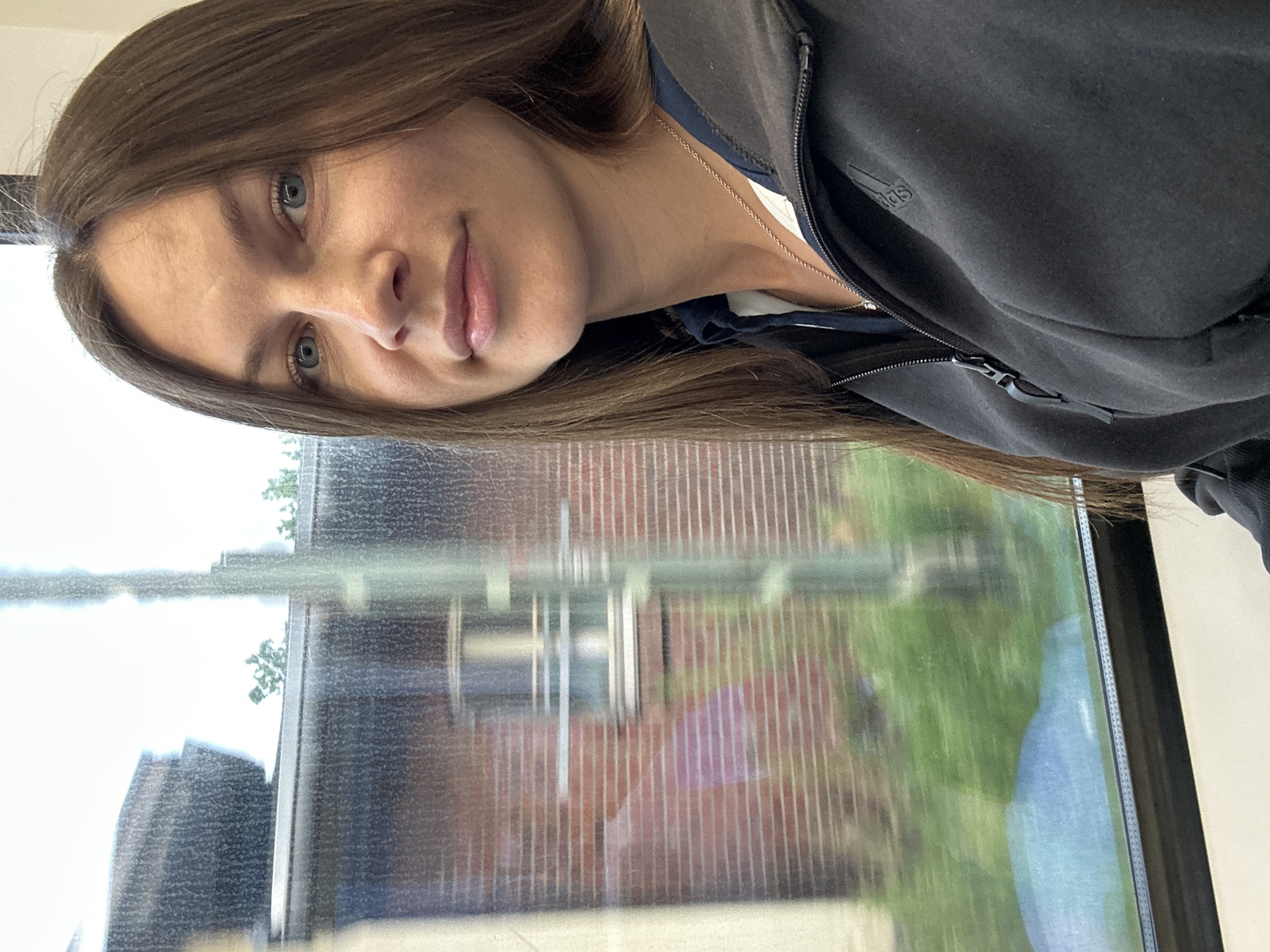 A portrait-style photo captures a young woman from the chest up, looking directly at the camera with a slight, soft smile. She has long, straight brown hair, parted on her left, framing her face. Her blue eyes are prominent, and she has fair skin. She is wearing a dark, possibly black or dark grey, zipped hoodie, under which a white garment and a thin silver chain necklace are visible. The "Adidas" logo is clearly discernible on the left side of her hoodie.
The woman appears to be seated or standing indoors, with a large window behind and to her side. The window glass is somewhat smudged or dirty and reflects elements of her hair and face. Through the window, an outdoor scene is visible, featuring green grass or foliage in the foreground, and a red brick building with vertical stripes or details further back. The sky above is bright, suggesting it is daytime, possibly overcast. The setting, according to the provided context, is Buxtehude, Germany. The overall mood of the image is calm and introspective.