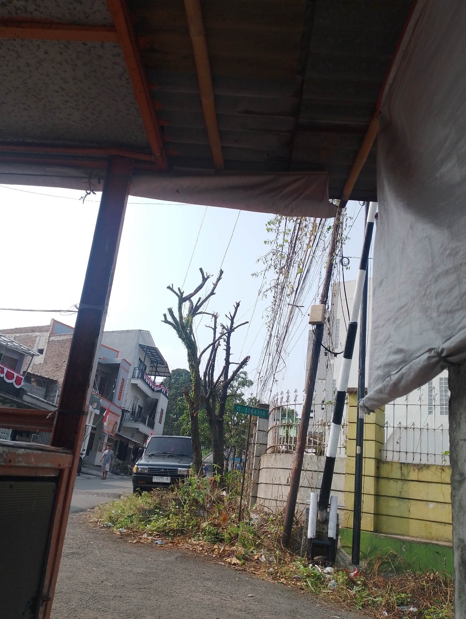 This is a street-level view of a residential area in Bogor, Indonesia. A dark-colored car is parked on the side of the asphalt road, which is lined with dry grass and foliage. In the background, several multi-story buildings with balconies and awnings are visible. The sky is clear and bright. A person is walking on the street in the middle distance. Utility poles with many wires are also present. A green street sign is partially visible on the left, indicating a street name. The foreground is dominated by the underside of a canopy or shelter with an orange metal frame and a grey roof. A white cloth is draped on the right side of the frame.
