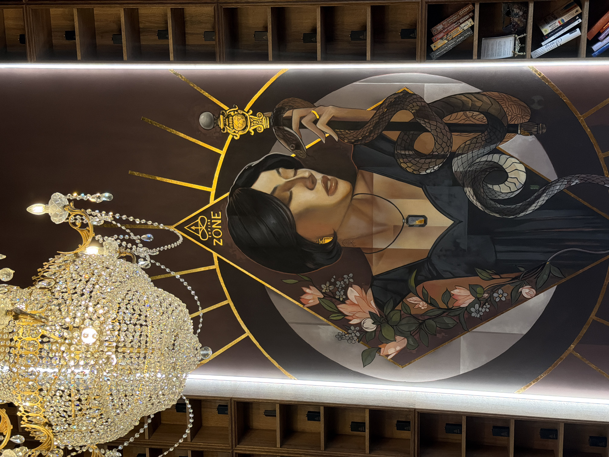 The image depicts a decorated ceiling with built-in shelves, likely in an upscale interior space. A large, ornate crystal chandelier hangs in the lower left of the frame. The ceiling mural features a woman with dark hair, eyes closed, possibly reclining. A snake is draped around her arm and hand, which holds a scepter or staff. Flowers are incorporated into the design near her lower body. There is text reading "EMPIRE ZONE" within the design. The scene is illuminated by a horizontal strip of light along the ceiling. The location might be in Moscow, Russia. Shelves containing books and other items are visible along the upper and lower edges of the frame.