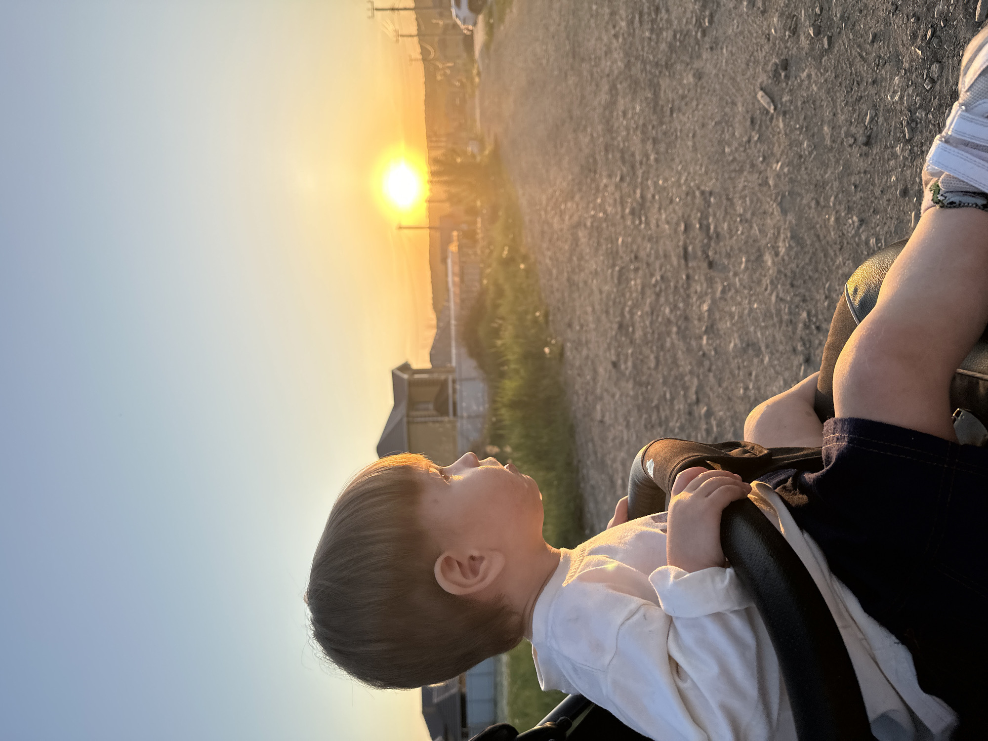 A low-angle shot shows a young child seated in a stroller, gazing towards a vibrant sunset. The child has short brown hair, fair skin, and wears a long-sleeved white shirt and dark shorts. The stroller’s black frame is visible. In the background, there's a dirt road and glimpses of houses, structures, and utility poles. The sun is a bright, circular orb, emitting a warm, golden light. Based on the location of Эстачи, Russia, and the presence of houses and roads, the environment appears to be a suburban or rural setting.