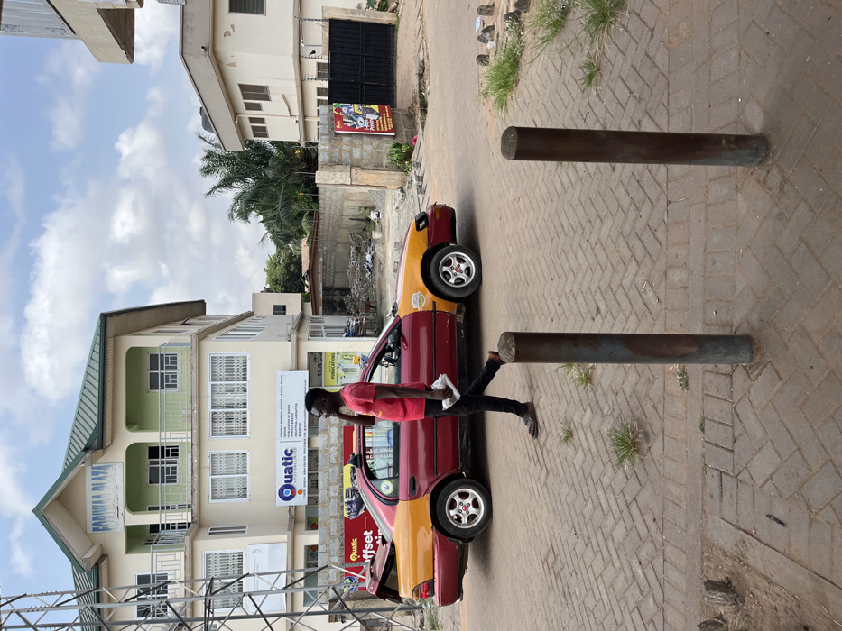 The image depicts an outdoor street scene within Adenta Municipality, Ghana, under a partly cloudy sky. In the foreground, two dark metal bollards are embedded in a reddish-brown brick-paved walkway, with sparse grass growth between the bricks.
A male individual, dressed in a red short-sleeved t-shirt, dark trousers, and brown sandals, walks from left to right on the paved walkway. He holds a white object in his left hand and a dark mobile phone to his right ear, appearing to be engaged in a phone conversation.
Behind the walking individual, a two-toned sedan vehicle, maroon on the lower body and yellow on the upper rear and front, is visible. The vehicle's trunk is partially open, and it features chrome hubcaps with red details.
In the background to the left, a multi-story building identified as "FODIWA PLAZA" stands, featuring a green sloped roof, cream-colored walls, and windows secured by white grilles. Ground-floor signage indicates businesses such as "Quatic Solutions Ltd" and "Offset Printing."
Adjacent to the FODIWA PLAZA, an open lot containing construction debris and rubble is visible, enclosed by a low concrete wall. Further right, a tall palm tree and another light-colored building with a dark gate are present. A vertical poster is affixed to a concrete pillar near the entrance of this building. The street surface beneath the vehicle appears to be asphalt.