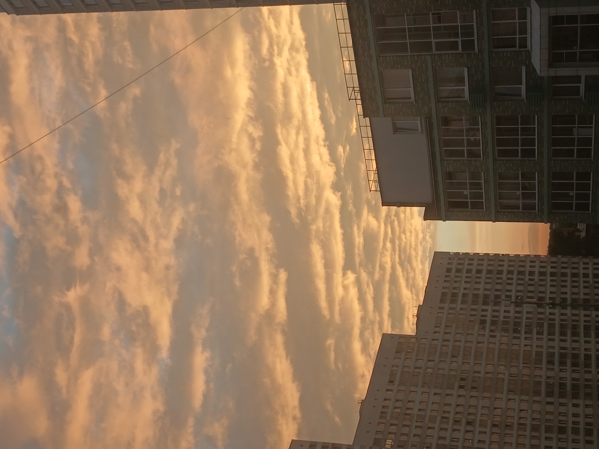 The image captures a view of buildings against a backdrop of a sunset sky filled with orange and yellow clouds. The buildings appear to be residential, with multiple windows visible on their facades. One building on the left is tall and mostly white, with a grid-like pattern of windows. Another building on the right is shorter, constructed with green bricks, and features larger windows, some with balconies. A thin wire stretches diagonally across the sky, likely an electrical or communication line. The scene is set in a city environment, and the overall impression is that of an urban landscape at dusk.
