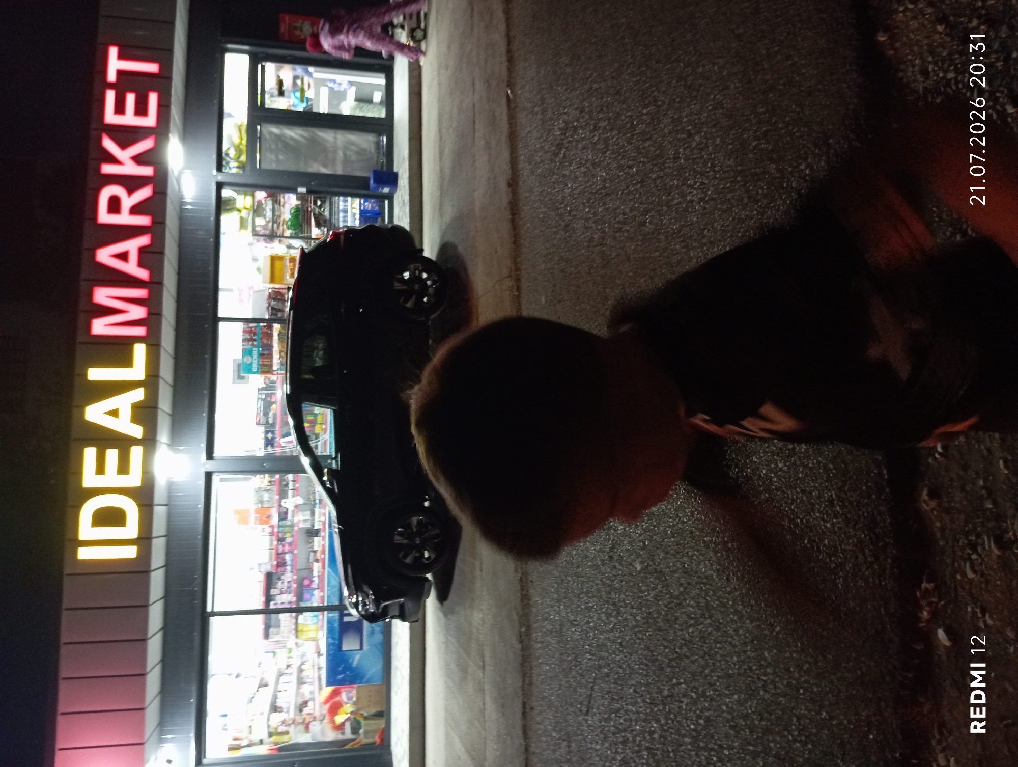 A nighttime street-level shot captures the facade of a "IDEAL MARKET" convenience store in Bekabad, Uzbekistan. The store's brightly lit sign, with yellow and red neon lettering, stands out against the dark exterior. Through the large glass windows, shelves stocked with various products are visible. In front of the store, a dark-colored SUV is parked, its headlights off. To the right of the store entrance, a person wearing a pink patterned outfit walks away from the camera, pushing a small cart. In the foreground, a young child's head and shoulders are visible, facing to the right and slightly out of focus. The ground in front of the store and in the foreground appears to be asphalt or a similar paved surface, wet from recent rain or moisture. The bottom left corner of the image displays the text "REDMI 12", and the bottom right corner shows the date and time "21.07.2026 20:31".