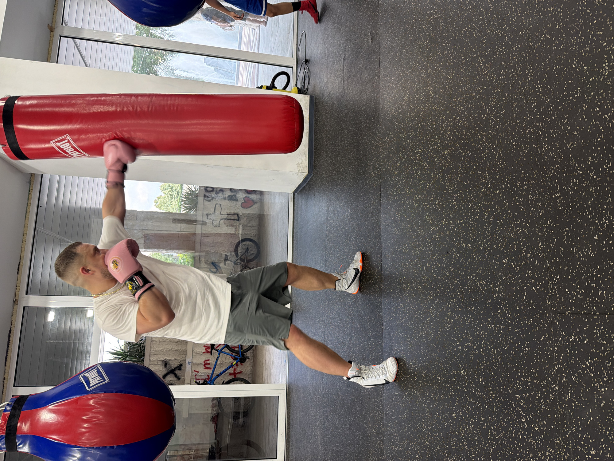 A man is training in a gym in Blagoevgrad, Bulgaria. He is wearing a white t-shirt, grey shorts, and white and orange boxing shoes. The man is punching a large red punching bag with pink boxing gloves on. To his left, there is a blue and red punching bag. In the background, there are large windows looking out onto a sunny day. There is also a bicycle and some graffiti on the wall. The floor of the gym is black and speckled.