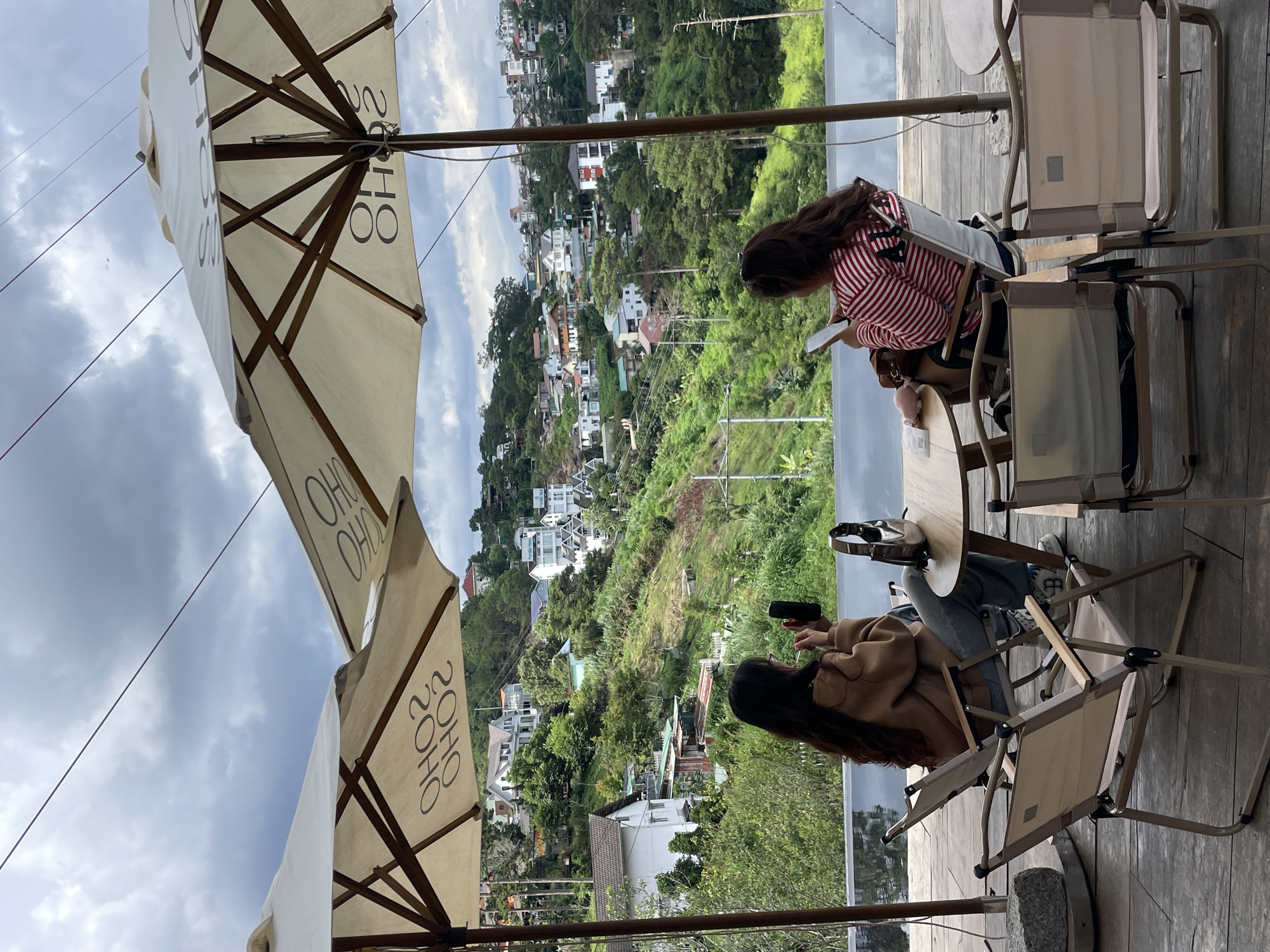 Two women are seated at a table outdoors, under large umbrellas, facing a hillside covered in houses and trees. The woman on the left, wearing a brown sweater and jeans, is holding a smartphone and appears to be taking a photo. The woman on the right, wearing a red and white striped shirt, is looking at her smartphone. Two folding chairs are in the foreground, and the table between the women has a handbag and a phone on it. The environment suggests a cafe or restaurant with a view in Da Lat, Vietnam, a city known for its hilly terrain and European-style architecture. The sky is cloudy.
