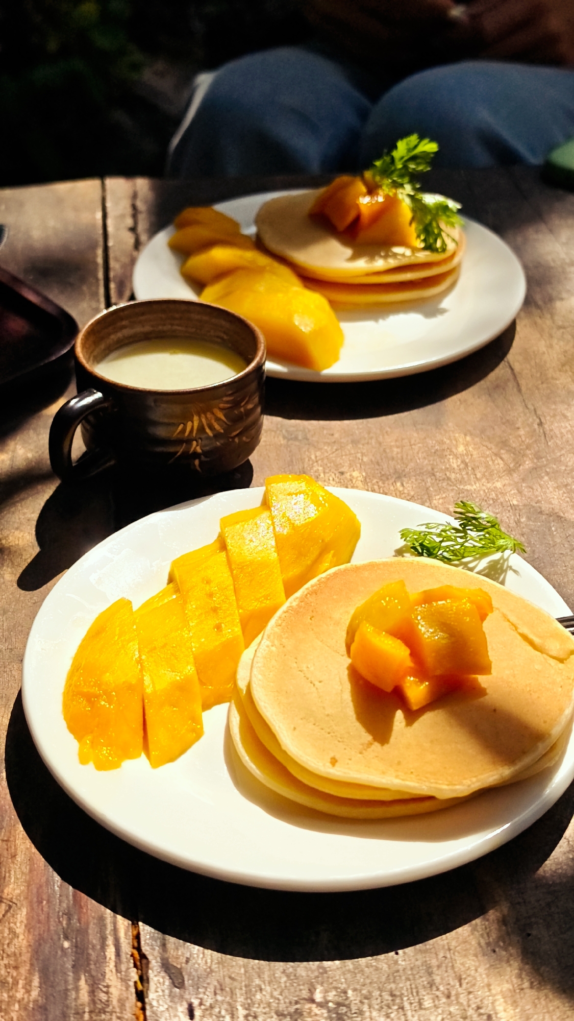 On a weathered wooden surface in Lũng Cú Commune, Vietnam, two plates hold pancakes topped with diced mango and sprigs of herbs. One plate is positioned closer to the viewer, showcasing a stack of pancakes, sliced mango, and green herbs. Nearby, a dark brown cup contains a creamy beverage. The second plate, slightly blurred in the background, mirrors the first with pancakes, mango, and herbs. Above the second plate, a person's legs are visible, suggesting they are seated at the table. The scene is brightly lit, highlighting the golden hues of the mango and pancakes against the dark wooden surface. Shadows indicate a strong light source from above.