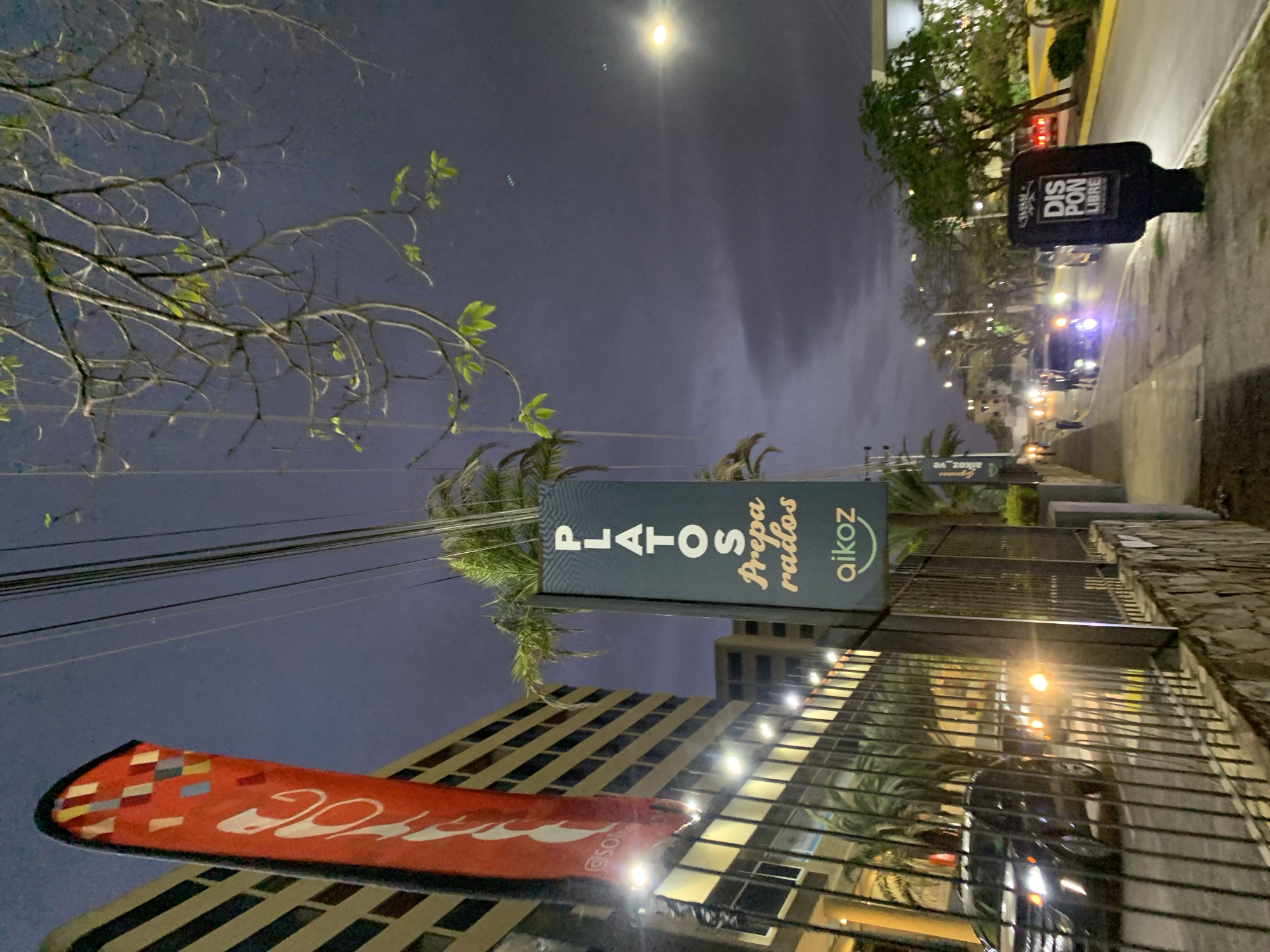 This media file captures a nighttime street scene, likely in an urban or commercial area of Pampatar, Venezuela. The sky is dark, illuminated by streetlights and a prominent, bright light source high above.
The foreground features a street with several cars, indicated by their illuminated headlights and reflections. To the right, a sidewalk runs alongside a building with a textured, possibly stone, facade and a metal railing. Further along the street, a multi-story building with large, reflective glass windows reflects the artificial lights of the surroundings. Another building with distinctive horizontal striped patterns is also visible.
Overhead, a network of power lines crisscrosses the dark sky. Various trees are present, including branches with a few sparse green leaves, and several tall palm trees that stand out against the night.
Several signs are visible:
1. A black, upright sign on the sidewalk reads "DIS PON LIBRE" in white text, likely indicating "DISPONIBLE" (available).
2. A large, rectangular sign, oriented vertically, displays "PLATOS Preparados" (prepared dishes) in white text and "aikoz" as a brand logo in orange on a dark blue background with a subtle pattern.
3. In the lower left, a red banner or flag, featuring a colorful checkered pattern on its left edge, shows white script text that is partially visible and appears to be a brand logo, possibly with a social media handle "@SO..." underneath.
The scene depicts a typical evening environment with artificial lighting defining the space, suggesting a commercial or residential zone where businesses are operating. No specific human activity is clearly visible, but the presence of cars and illuminated signs implies regular urban life.