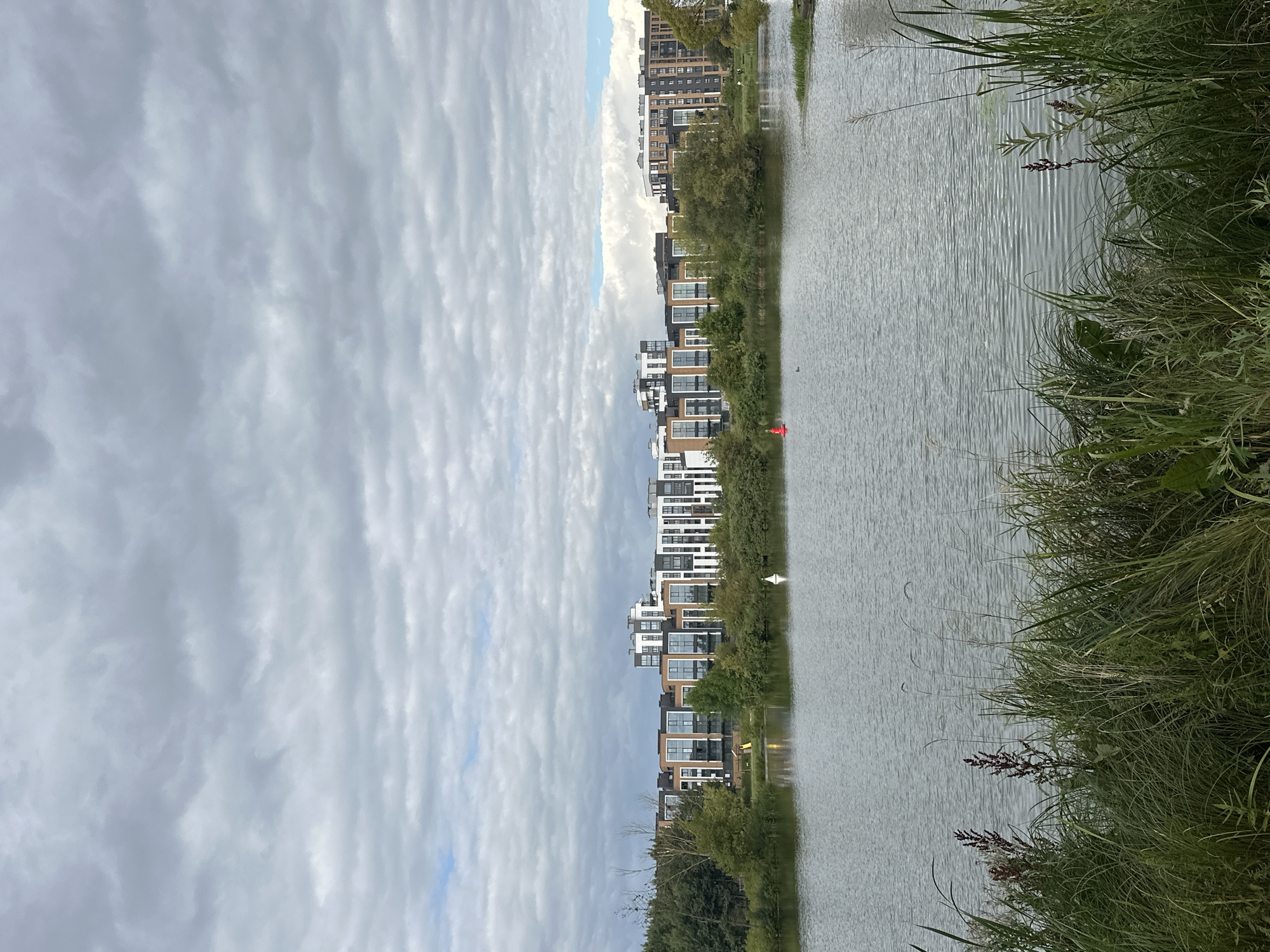 The image shows a lake with a modern apartment complex in the background. The sky is cloudy and overcast. In the foreground, there is tall grass and reeds growing along the edge of the water. In the middle ground, on the lake, there is a white buoy and a red buoy. A single white bird is visible near the white buoy. The apartment complex is multi-story with a mix of white, brown, and dark gray facades, and many windows. Trees are interspersed between the apartments and along the shoreline. The overall scene depicts a calm, urban waterscape with residential development. The location is identified as Minsk, Belarus.