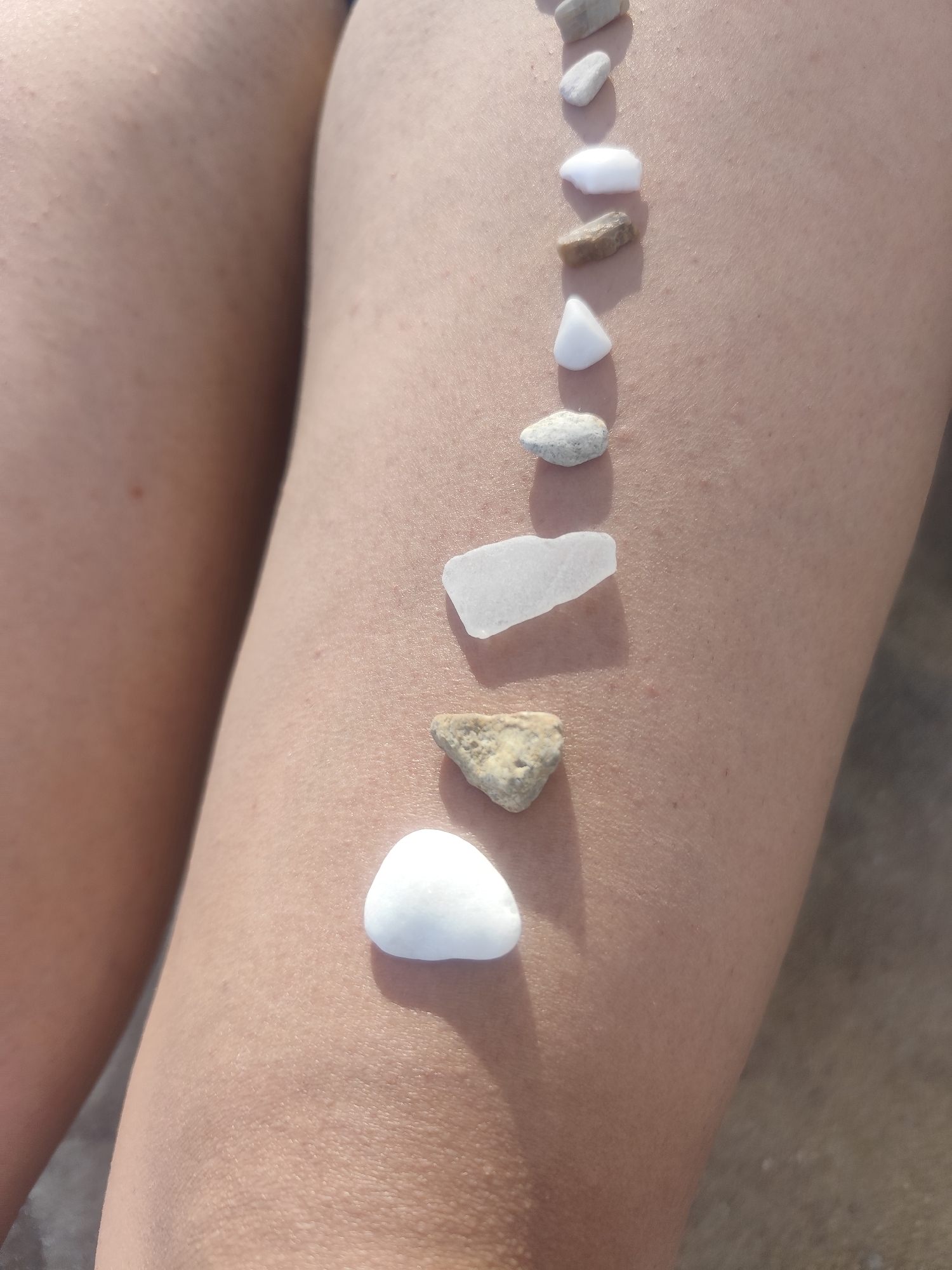 A close-up, high-angle shot shows a line of smooth, pale-colored pebbles and sea glass arranged vertically on the bare thigh of a person. The skin appears tanned and lightly textured. The pebbles vary in size and shape, with some being rounded and others more angular. The sea glass piece is translucent and irregular. The background is blurred, suggesting an outdoor environment, possibly a beach in Kavala, Greece, given the context. The lighting suggests it is daytime, with a soft shadow cast by each object.