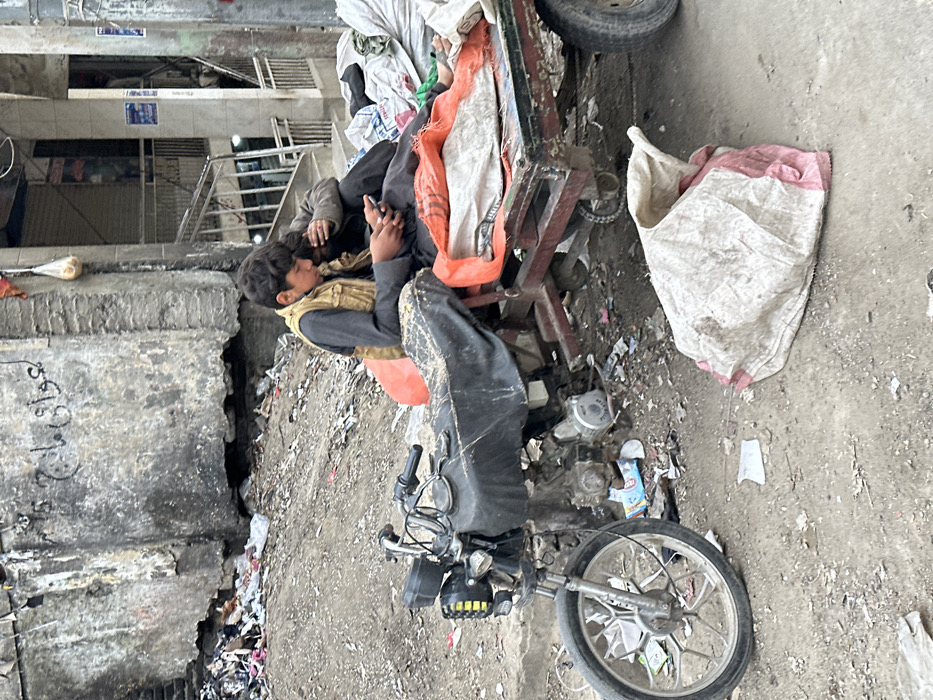 This image depicts an outdoor urban scene in Quetta City Tehsil, Pakistan, characterized by squalor and daily activity.
In the foreground, a modified black motorcycle with a cargo trailer attached to its rear is parked on a dirt and gravel surface. The motorcycle seat is covered with black material, and an orange tarp or blanket is visible on the trailer bed, along with several white and orange sacks. Two individuals, both appearing to be young males, are seated on the cargo trailer. The individual on the left, wearing a beige vest over a dark long-sleeved shirt and dark pants, is looking down at a black smartphone held in his hands. His left foot is bare and resting on the trailer bed. The individual on the right, also in dark clothing, is leaning against the first person, his face mostly obscured by his arm or hair. Both appear to be focused on the smartphone.
The ground around the motorcycle and trailer is unpaved, consisting of packed dirt and loose gravel, scattered with various pieces of trash, including plastic wrappers, paper, and debris. To the right of the motorcycle, a large, partly empty white and red striped sack lies on the ground.
In the mid-ground, behind the individuals and vehicle, a concrete or stucco wall of a building is visible. This wall is heavily stained, weathered, and shows signs of damage, with debris piled against its base. Graffiti in Urdu script is discernible on the upper portion of the wall. To the right of this wall, a multi-story building with a metal railing staircase leading upwards is present. The lower section of this building appears to house shops or stalls, with some items hanging outside. Overhead, wires are visible. The overall environment suggests a neglected urban area.
