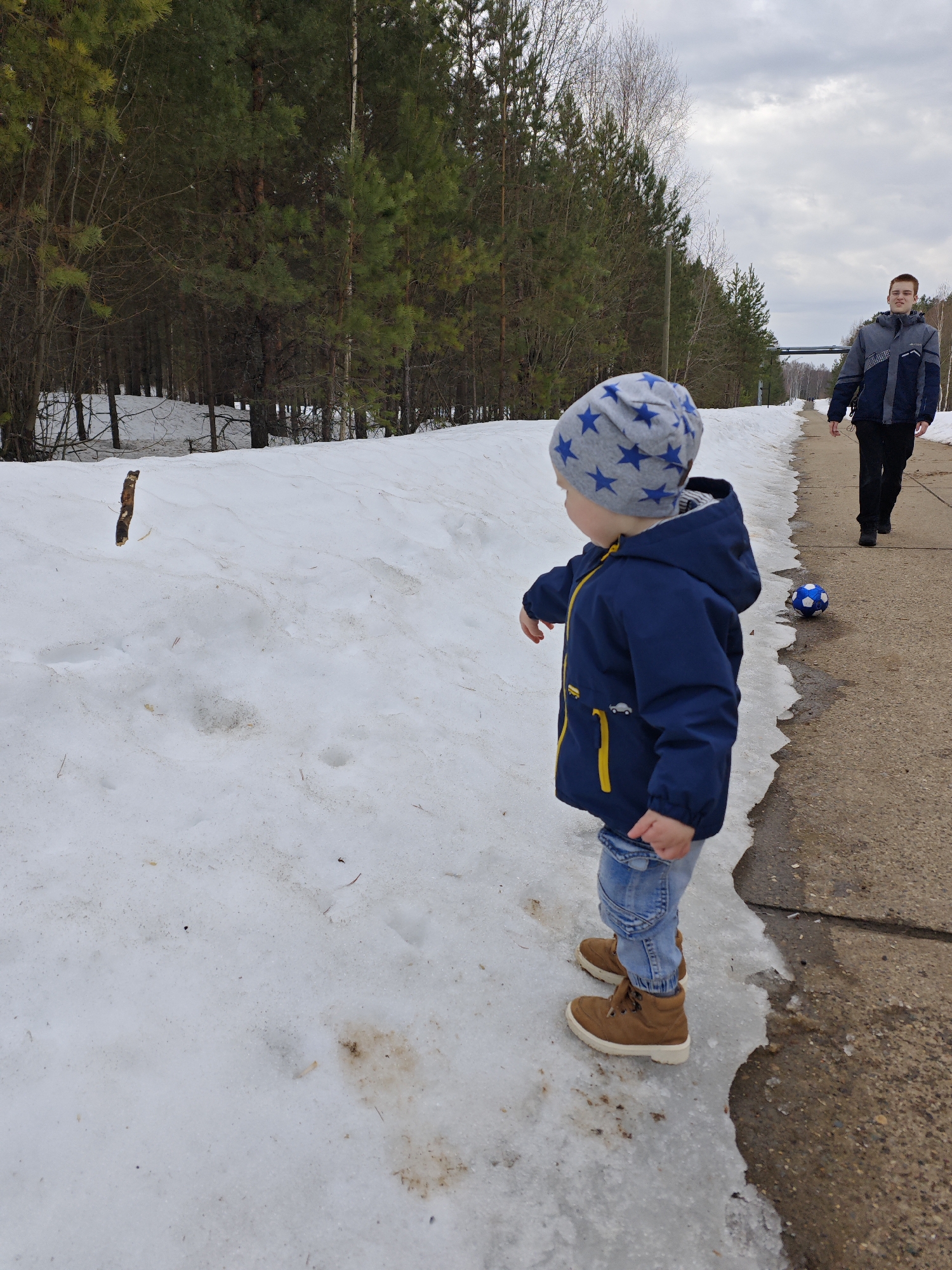 The image depicts an outdoor scene in what appears to be early spring in Марковский, Russia. A young child, wearing a blue jacket with yellow trim, light blue jeans, and brown boots, stands on a patch of melting snow and ice. The child is also wearing a gray hat with blue stars. To the right of the child, a blue and white soccer ball sits on a concrete path. Further down the path, an adult male is walking away from the child. The adult is wearing a gray and blue jacket and black pants. The background shows a snow-covered landscape with a line of evergreen trees and a cloudy sky.