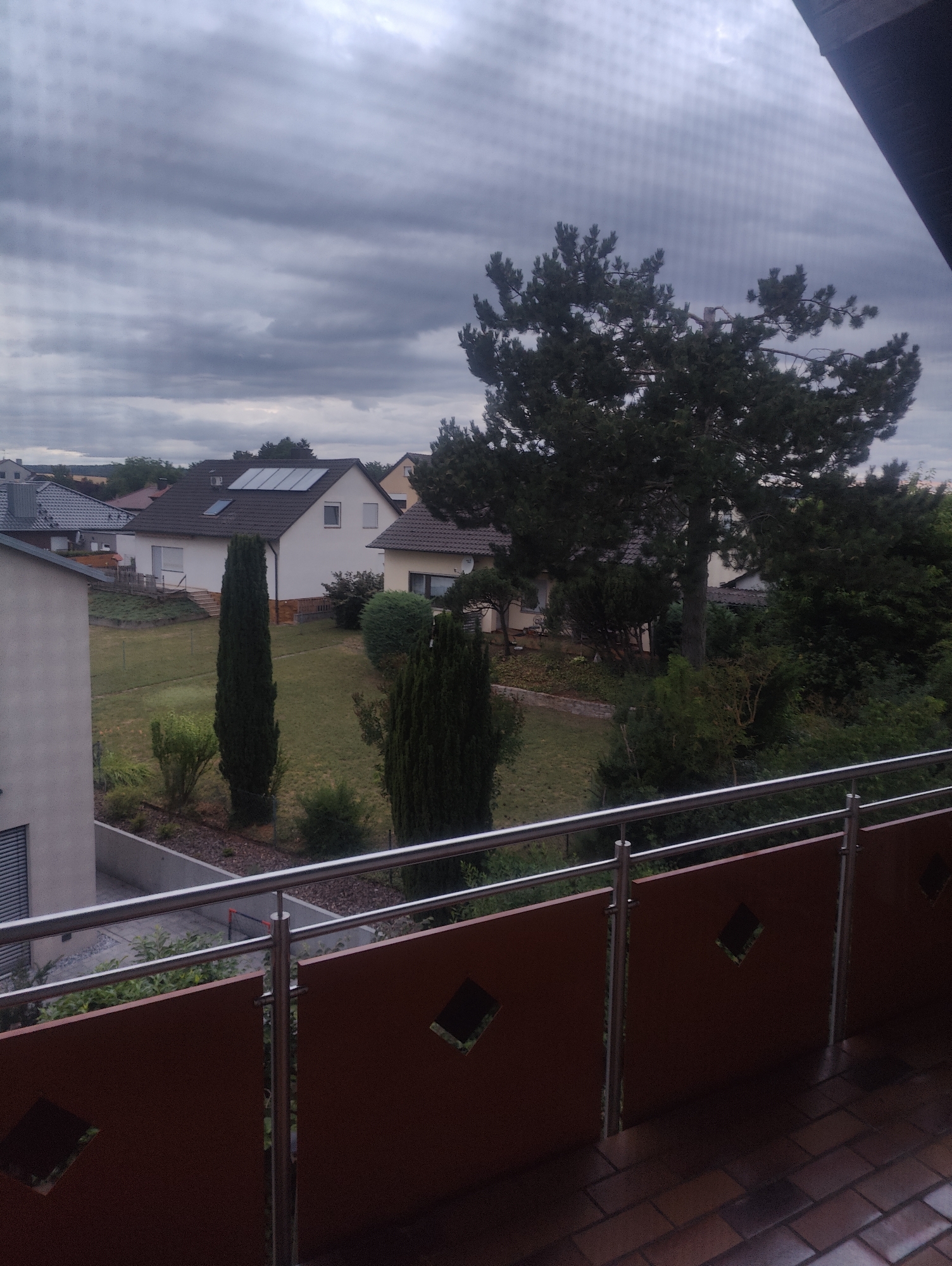 This is an outdoor, medium shot of a residential area under a cloudy sky, taken from a balcony. In the foreground, a brown balcony railing with diamond-shaped cutouts and a metal handrail is visible. Beyond the railing, a small lawn and garden area are present with several houses and trees. One house in the middle distance has solar panels on its roof. The sky is overcast with dark grey clouds.