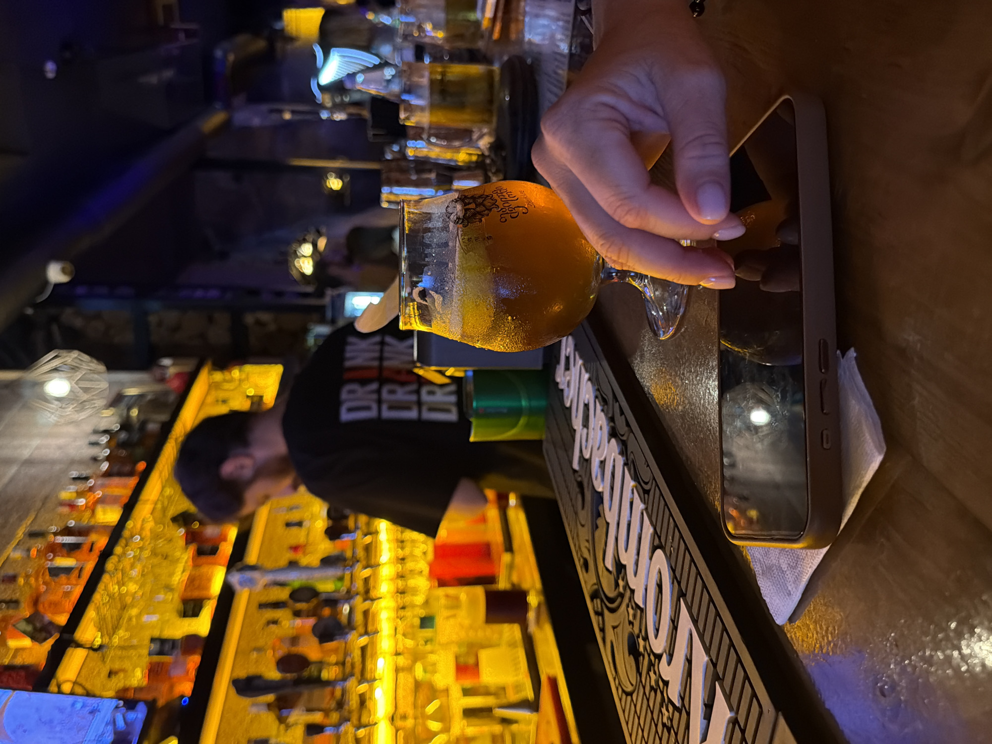 The image depicts a dimly lit bar environment, likely in Batumi, Georgia, with a focus on a glass of amber-colored liquid, possibly beer, resting on a wooden counter. A person's hand, with manicured fingernails, is visible, seemingly interacting with a smartphone placed on a white napkin. In the background, a bartender in a black t-shirt with white lettering that reads "DRINK DRINK DRINK" is partially visible, facing away from the camera. Behind the bartender, shelves are stocked with numerous bottles of alcohol, illuminated by warm, golden lights. Various taps for beverages are also visible. The overall atmosphere suggests a relaxed setting, perhaps a tourist-oriented establishment, given the potential location in Batumi. The lighting creates reflections on the counter and the smartphone screen, adding to the ambiance.