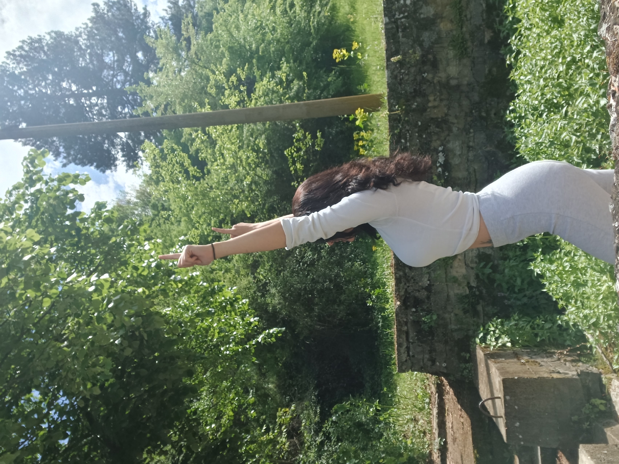 A woman is standing on a stone structure, potentially ruins, in what appears to be a natural, wooded area. She is wearing a light-colored long-sleeved shirt and gray leggings. She is facing towards the left of the frame with her arms extended straight out, pointing with her index fingers. The background consists of dense green trees and foliage, with a glimpse of sky and a wooden beam visible. The photograph may be taken in Gorna Verenitsa, Bulgaria based on context.