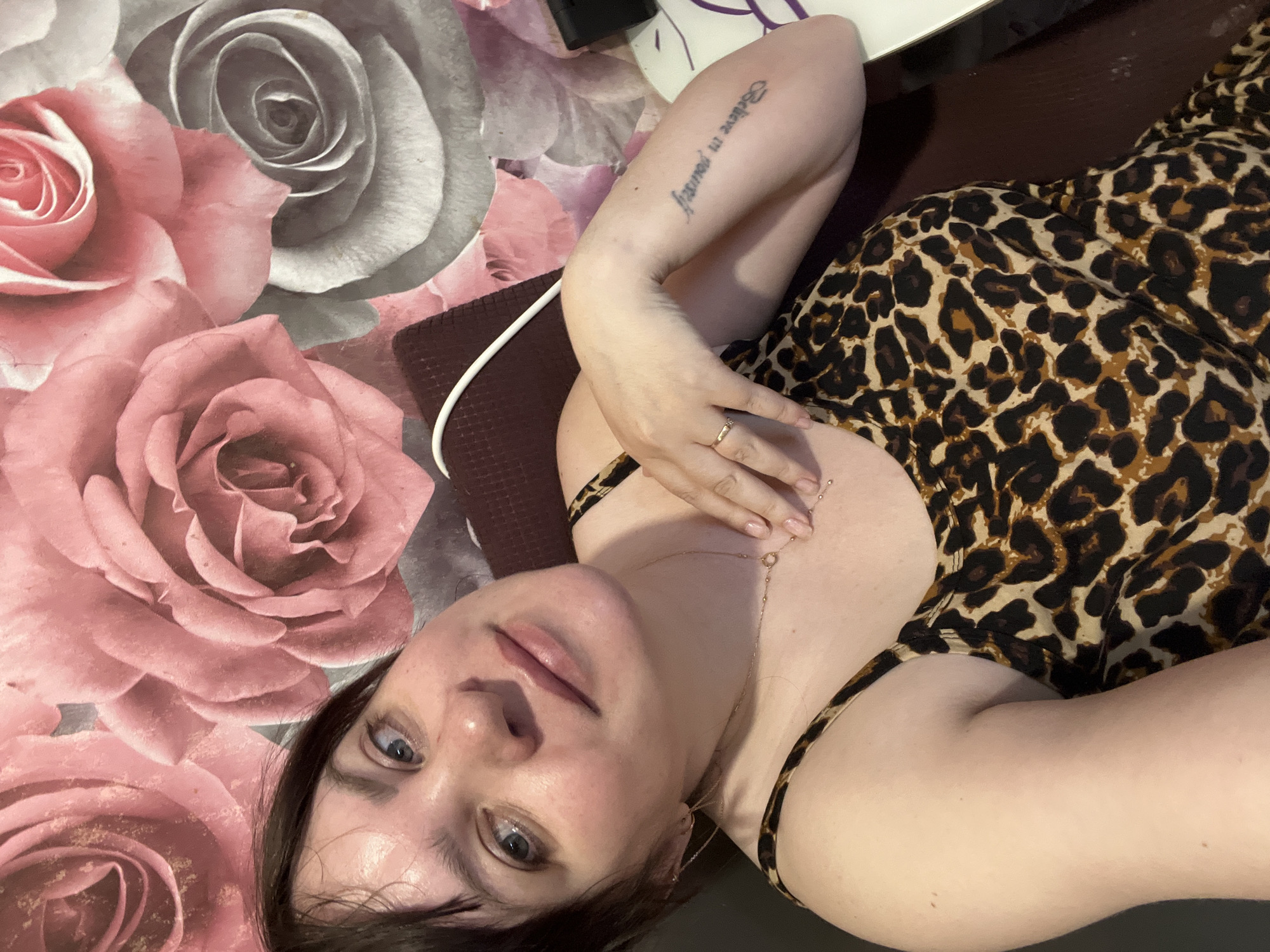 A young adult female lies on a patterned surface, likely fabric or wallpaper, featuring large pink and gray roses. She is wearing a leopard print dress and a gold chain necklace. Her left arm is raised, revealing a tattoo that reads "Believe in yourself." She has a ring on her finger. The location is indeterminable from the image; there is no information to connect the image to the city of Noviy, Russia.