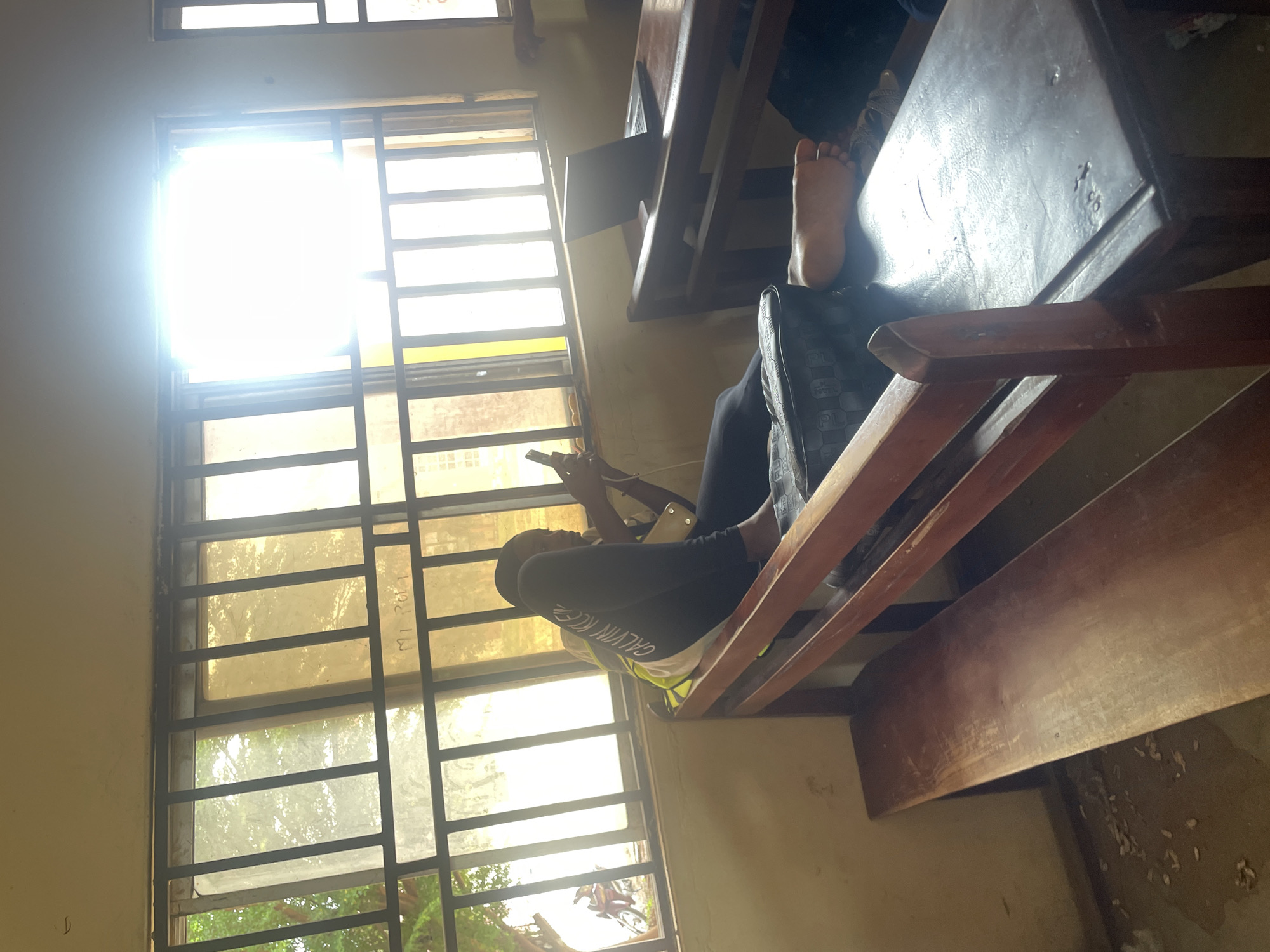 A person is sitting at a desk in a classroom. The person is looking down at a phone in their hands. The person is wearing a black long-sleeved shirt with white lettering on it, and white shorts. Their feet are resting on another desk. The classroom appears to be old, with wooden desks and chairs. The windows are covered with metal bars. Sunlight is streaming through the windows, creating a bright glare. There are trees visible outside the window. The scene is likely taking place during the daytime. The lettering on the shirt appears to say "CALVIN KLEIN".