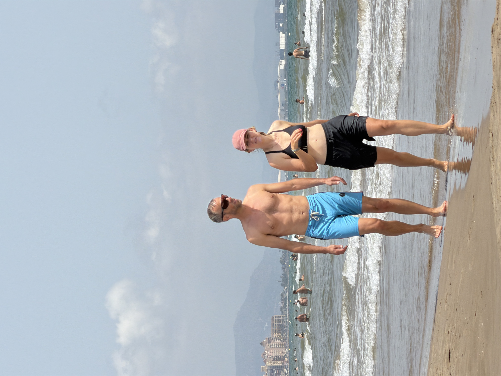 The media file shows two people on a beach with the ocean and a cityscape in the background. The image is rotated 90 degrees counter-clockwise. In the foreground, a man wearing sunglasses and blue swim trunks stands with his feet in the shallow water. He is looking towards the ocean. To his left and slightly behind him, a woman wearing a pink cap and a black bikini top and black shorts is lying on the wet sand, propped up on her hands. She is looking towards the camera. The ocean has waves and several other people are visible in the water and on the beach in the distance. A hazy cityscape with buildings is visible on the shore, with mountains behind it. The sky is overcast. The overall impression is a sunny day at the beach. There is no visible text.