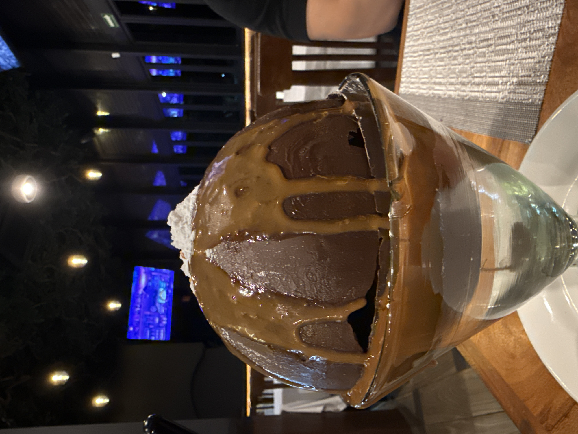 The image shows a large chocolate dessert in a glass, possibly a sundae or a similar layered dessert, with chocolate ice cream or mousse, whipped cream, and caramel sauce. The dessert is on a wooden table, next to a white plate. The background shows an outdoor setting with lights and possibly trees, with a building with lit windows and a television in the background. It appears to be nighttime. Based on prior information, the location is likely Mexico City, Mexico. A person is sitting at the table, only partially visible in the upper part of the frame. The scene depicts someone dining outdoors at a restaurant in Mexico City.