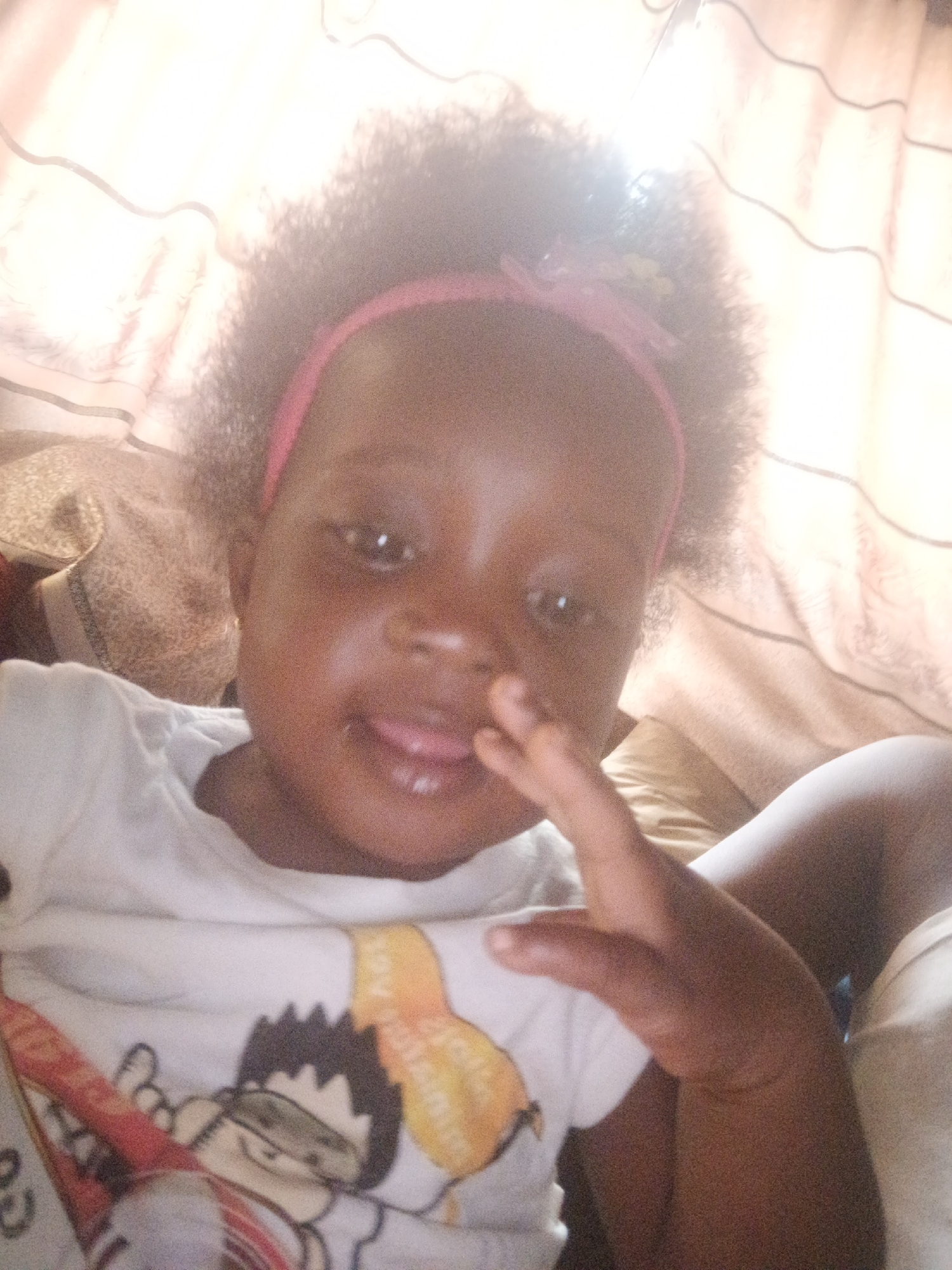 A close-up shot features a young child with dark skin, wearing a pink headband with a floral embellishment. The child is sticking their tongue out slightly and has their right hand raised, fingers partially curled. They are wearing a white t-shirt with a cartoon character design on it. The background includes light-colored curtains and a partially visible beige pillow or cushion. The image was likely taken indoors. There is no explicit information to place the location within Akwanga, Nigeria.