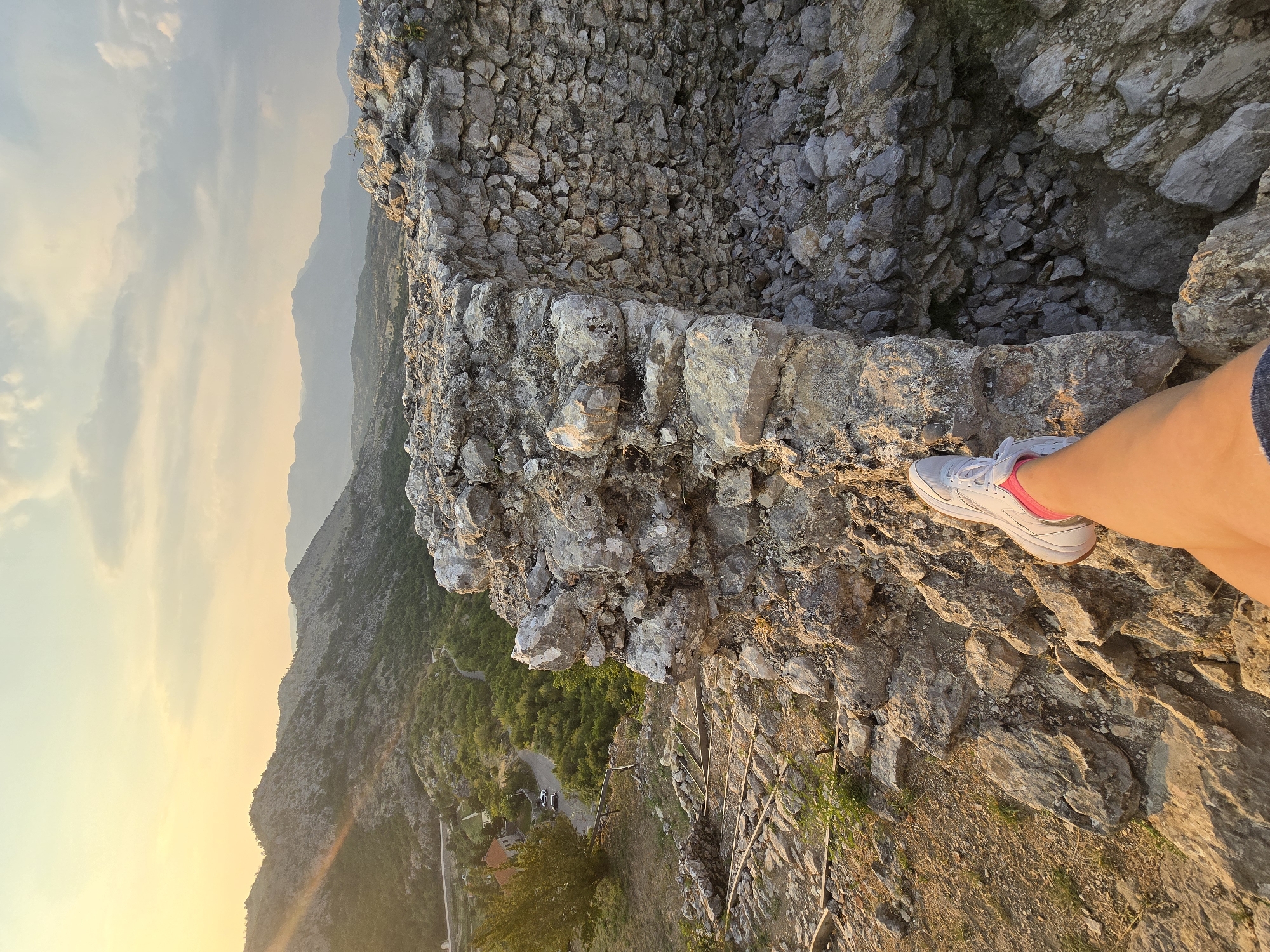 The image shows a first-person perspective of a person's leg, wearing white sneakers with pink accents and denim shorts, resting on the ancient stone ruins of Medun, Montenegro. The ruins, constructed from rough, grey stones, form a wall in the foreground. Beyond the ruins, a lush green valley unfolds, with a winding road and a red-roofed building visible in the distance. The background is dominated by rolling hills and distant mountains, bathed in the warm, golden light of a sunset. The sky is a mix of yellow and blue, with scattered clouds. The overall scene evokes a sense of historical exploration and scenic beauty.