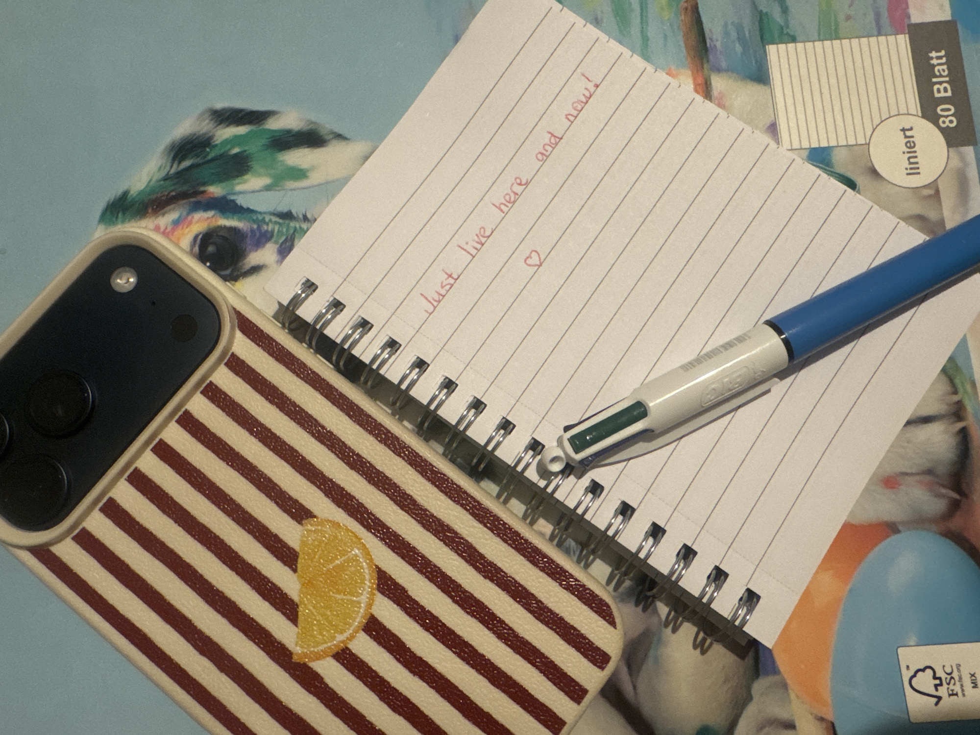 The image displays a close-up, overhead view of a collection of personal items arranged on a surface with a colorful, abstract pattern featuring a dog with vibrant markings. In the foreground, a spiral-bound notepad with lined paper is open, with red handwritten text that reads "Just live here and now!" accompanied by a small heart symbol. A blue and green multi-function pen rests on the notepad. Behind the notepad, a portion of a smartphone case is visible. This case has a cream-colored background with alternating maroon stripes and a small, embroidered slice of orange on its surface. The camera module of the smartphone is also visible at the top edge of the case. To the left of the notepad, a light blue plastic object with an FSC MIX certification label is partially visible, along with an orange rounded object. The overall scene appears to be a still life composition of everyday objects.