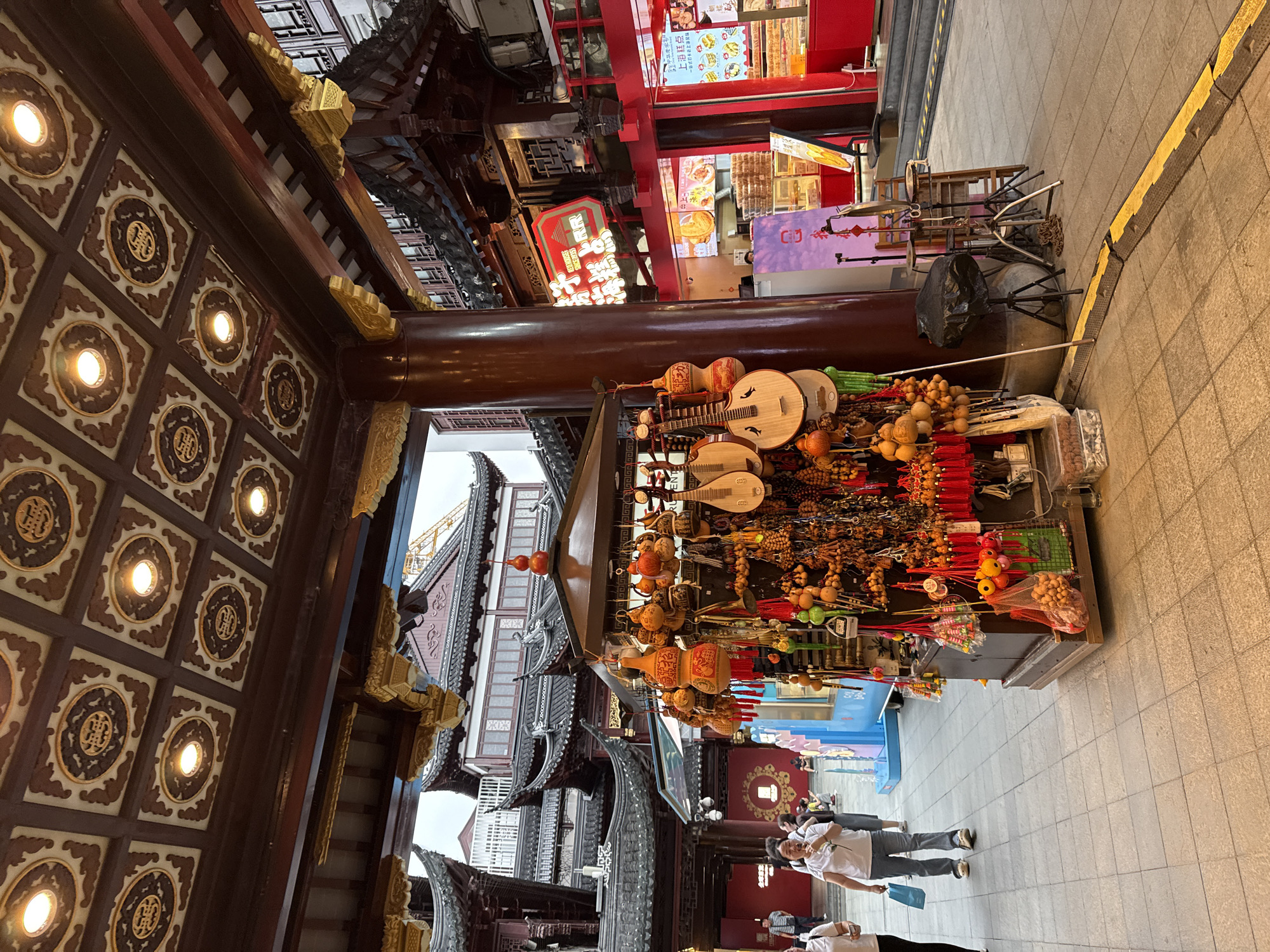 The image depicts a bustling street scene in Shanghai, China, specifically within the Yu Garden Bazaar, a historic tourist attraction known for its traditional architecture and vibrant market. The foreground is dominated by a stall overflowing with traditional Chinese handicrafts, including numerous small gourds, musical instruments resembling lutes, and red tassels, all suspended from the stall's canopy.
To the left, two women are walking on a tiled pathway. One woman, wearing a white t-shirt and blue jeans, is holding a white flower and a blue shopping bag. The other woman, in a white t-shirt and black pants, is walking ahead of her. The pathway is paved with square, light-colored tiles, and a yellow and black speed bump runs across it in the lower right quadrant.
The background reveals the distinctive architecture of the Yu Garden Bazaar. Traditional Chinese buildings with curved, dark tiled roofs and ornate wooden eaves are visible. A large, dark red pillar supports an overhanging structure, and the ceiling above it is intricately decorated with square panels featuring gold and dark brown motifs and illuminated by recessed lights.
Further back, shops with bright red facades line the street, displaying food items and promotional signage, including a neon sign with Chinese characters. A blue structure, possibly a play area or information kiosk, is also present. The overall impression is one of a lively, culturally rich urban environment.