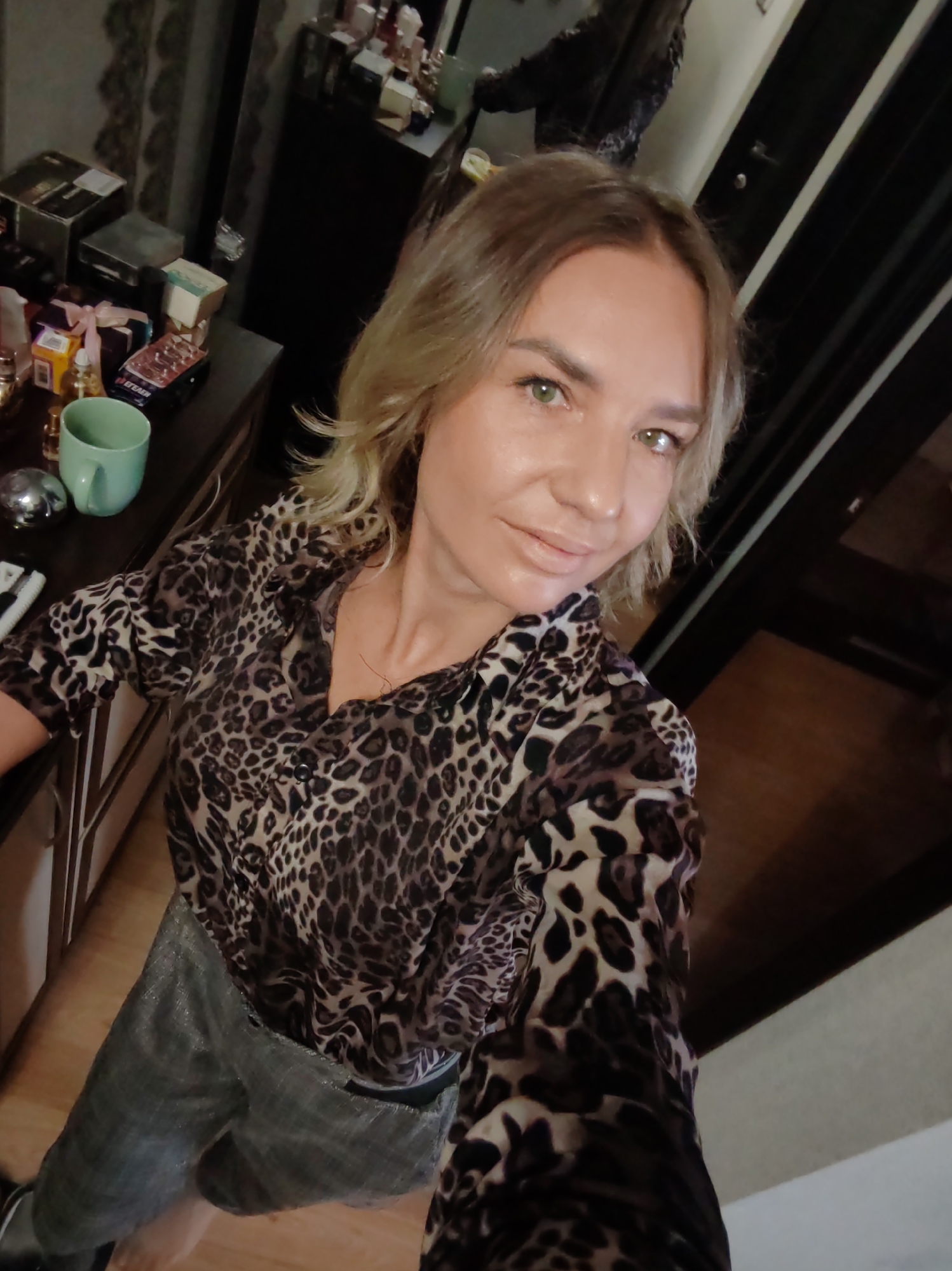 The image features a fair-skinned woman with shoulder-length, wavy blonde hair and light green eyes, taking a selfie indoors. She is wearing a V-neck, long-sleeved shirt with a brown and black leopard print pattern, and shimmery gray trousers. She looks directly at the camera with a subtle smile.
The setting appears to be a room within a private residence in Petropavlovsk-Kamchatsky, Russia. The background on the left shows a dark wooden dresser or shelf filled with various personal items. These include several perfume bottles, various cosmetic boxes (one of which has the partially visible Cyrillic text "БЕЛАЯ"), a light teal-colored mug, and a silver spherical ornament. A dark wooden doorframe is visible on the right side of the image, and a darker, possibly reflective surface or mirror stands behind the dresser. The artificial lighting suggests the photo was taken in the evening or at night.