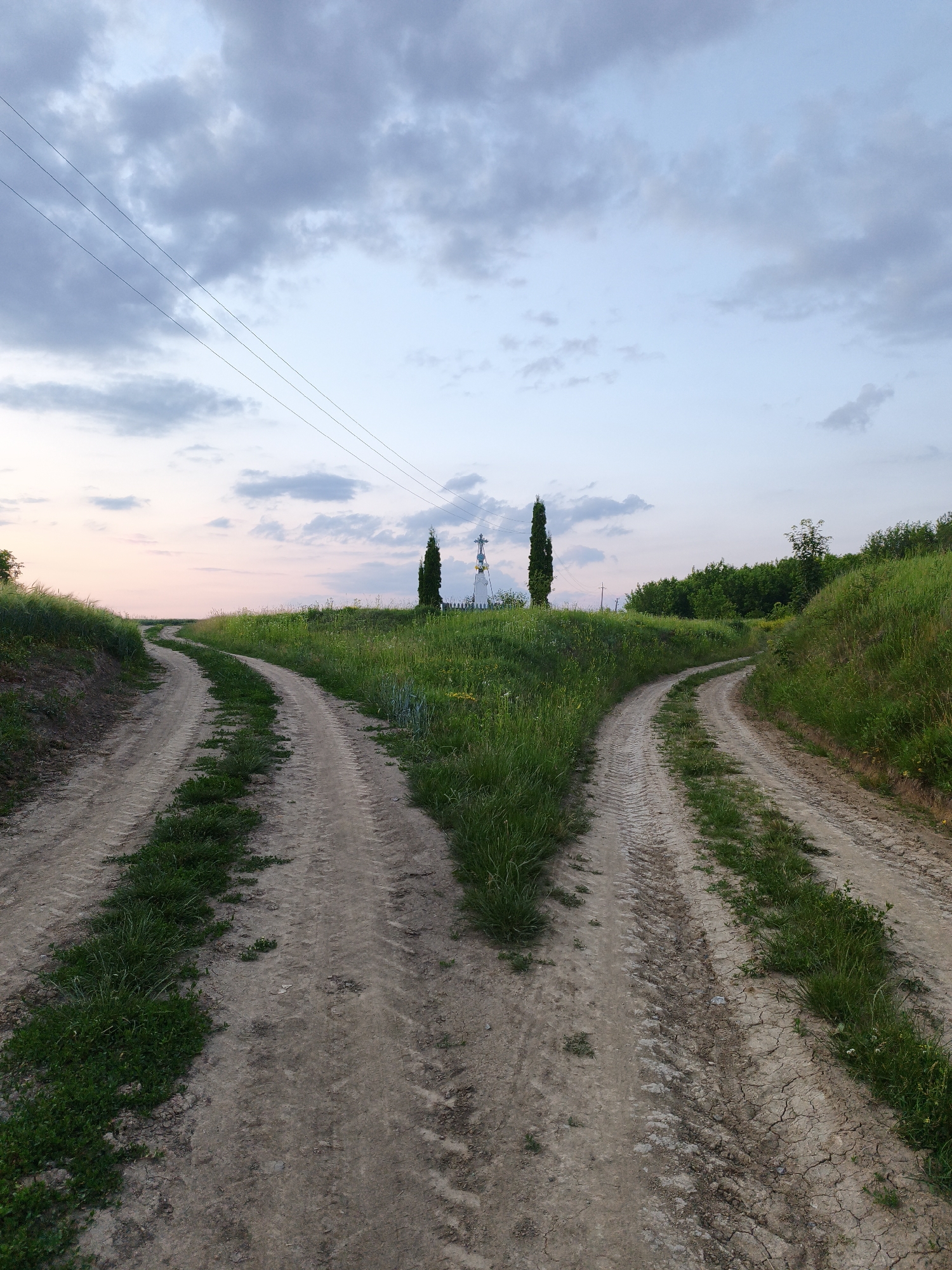 The image captures a fork in a dirt road in a rural landscape. The road, with visible tire tracks, splits into two paths that diverge as they ascend a grassy hill. The left path curves to the left, while the right path curves to the right. In the distance, atop the hill, a white cross or monument is visible, flanked by two tall, slender trees. Power lines stretch across the sky in the upper left portion of the frame. The sky is partly cloudy, with hues of blue and soft pink from what appears to be either sunrise or sunset. Lush green grass and wildflowers grow along the sides of the road and on the hills. The overall scene suggests a serene, natural setting.