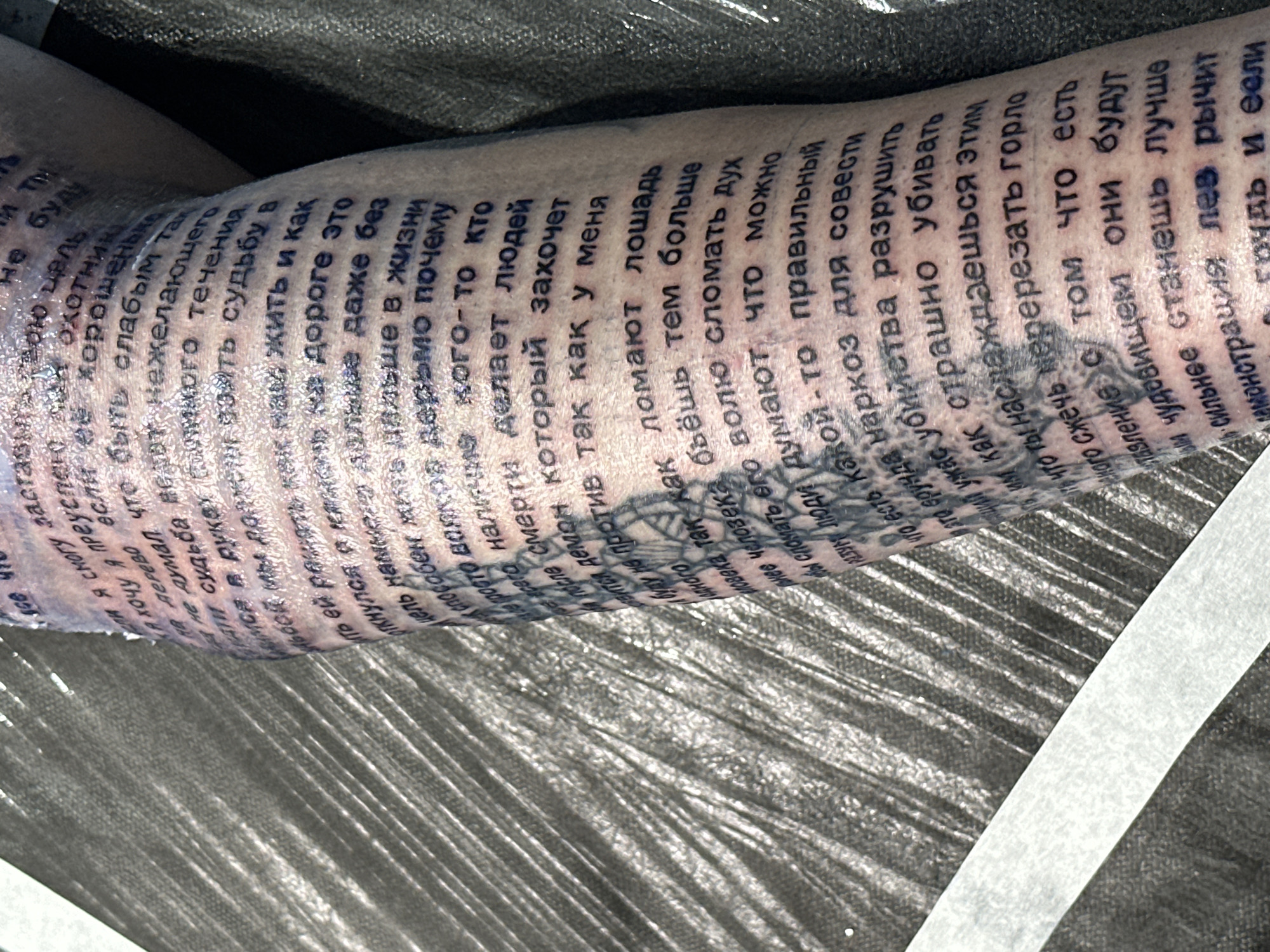 Here is a detailed description of the image:
The image shows a close-up of a person's arm that is covered in a tattoo. The tattoo is comprised of lines of Russian text, running along the length of the arm. The arm appears freshly tattooed, as it is covered in a clear protective wrap. The surface beneath the arm is a crinkled metallic silver color. A strip of what appears to be white medical tape is visible near the bottom right corner of the image. The lighting in the image is somewhat direct, causing some glare on the plastic wrap. The text is black and in a font that is dense and closely spaced.