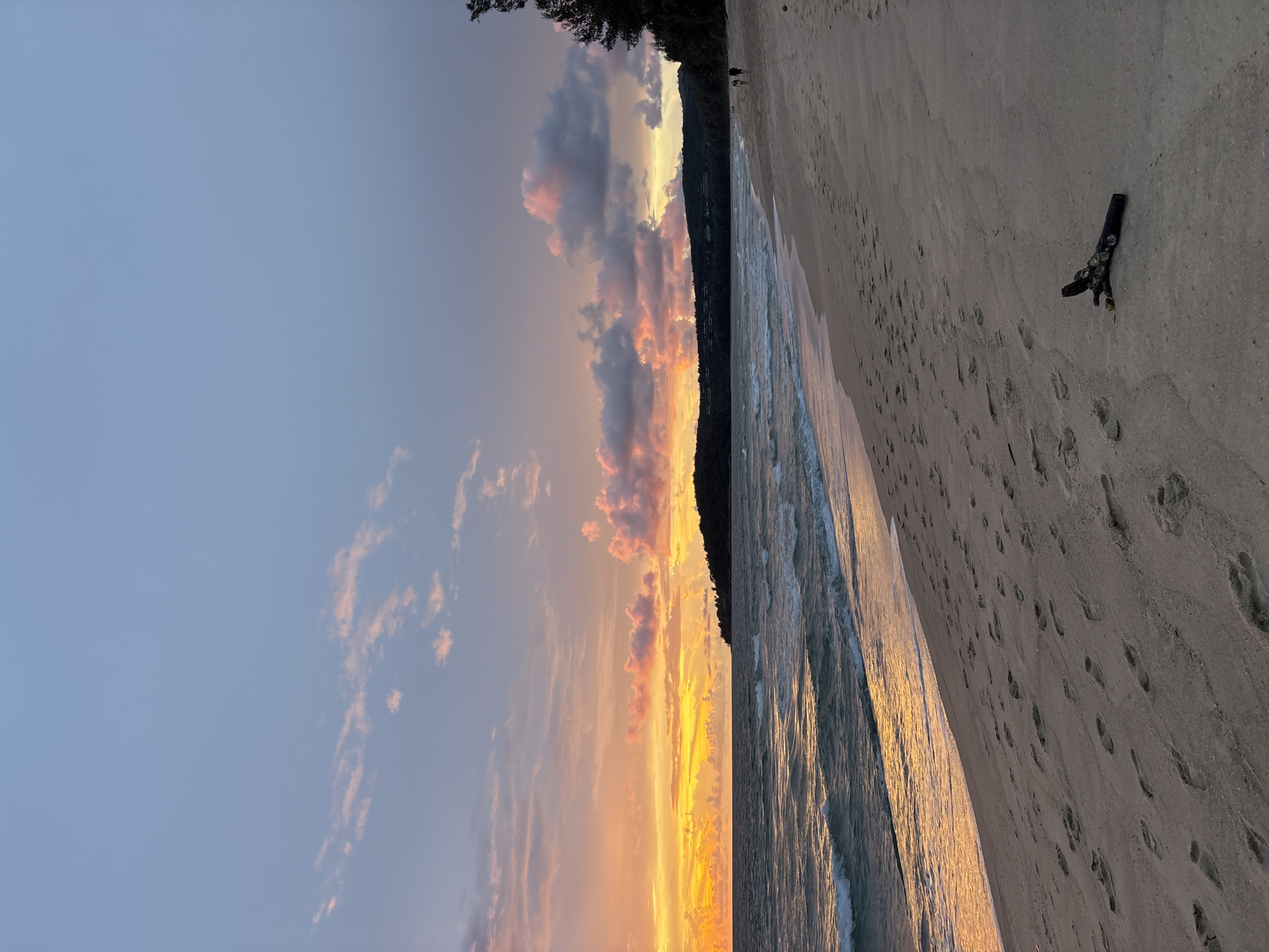 The image shows a beach at sunset in Choeng Thale, Thailand. The sky is a gradient of soft blues, pinks, and oranges, with scattered clouds illuminated by the setting sun. The ocean is calm, with gentle waves lapping at the shore, reflecting the warm colors of the sky. The sandy beach is marked with numerous footprints, suggesting recent activity. In the foreground, a piece of driftwood lies on the sand. In the distance, a line of trees forms a dark silhouette against the vibrant sky, and two figures are walking along the beach. The overall scene is serene and picturesque.