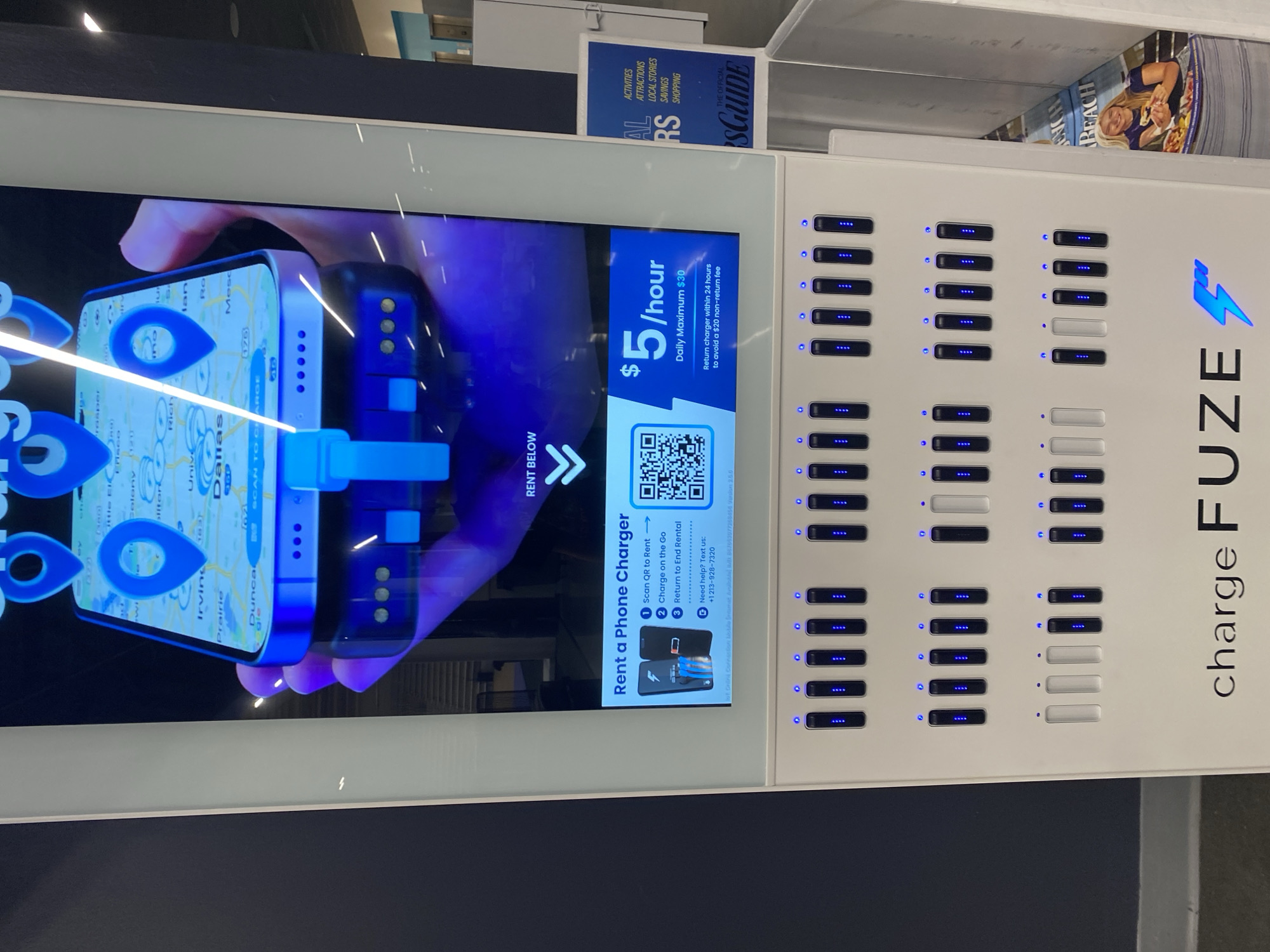 The image shows a ChargeFuze phone charging station located in Virginia Beach, United States of America. The charging station is white and features a digital screen displaying a map of Dallas, Texas with location pins. Below the screen, there is text explaining how to rent a phone charger, listing the cost as $5 per hour with a daily maximum of $30. There are multiple charging bays, most of which are occupied by dark-colored charging units with blue indicator lights. A few bays appear to be empty or occupied by white units. In the lower right corner, a magazine or brochure is visible with the word "BEACH" on it and a picture of a woman. The background is dark, suggesting an indoor environment, possibly a public space like an airport or mall.