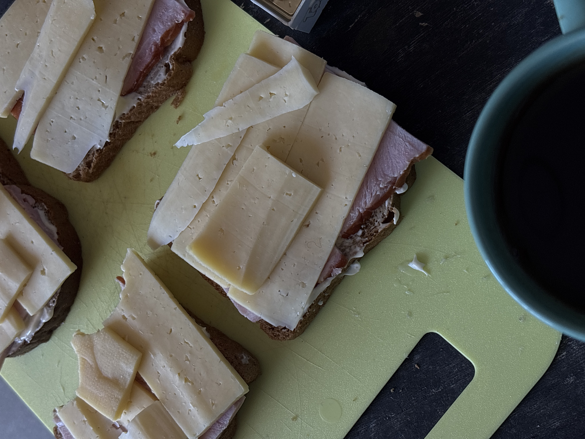A top-down view reveals a yellow cutting board holding open-faced sandwiches with sliced cheese, ham, and bread. A mug containing dark liquid is partially visible on the right. A small box is also present on the board. The location cannot be determined from this image.