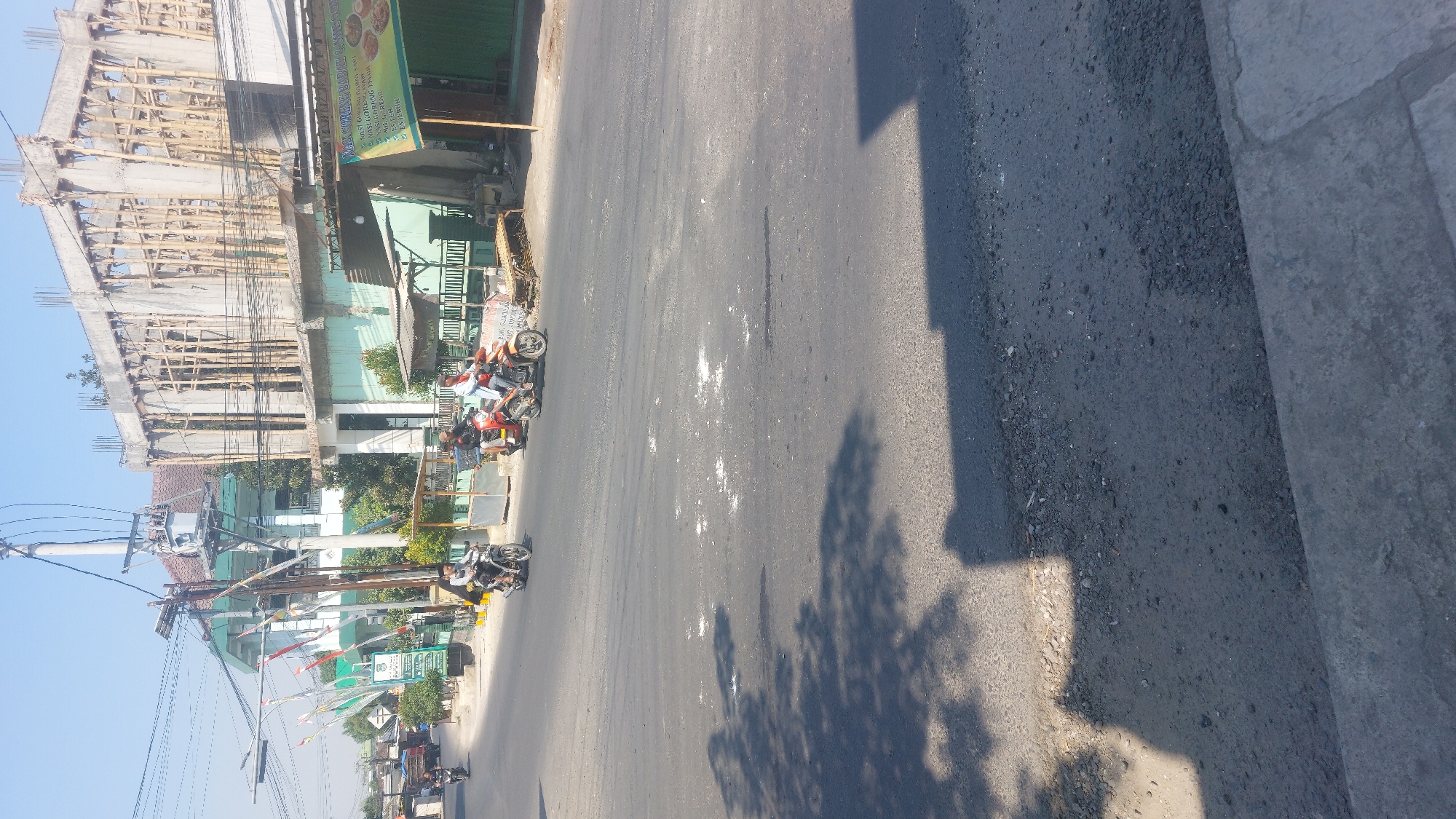 The image depicts a street scene in Sedan, Indonesia, under a clear sky. In the foreground, the asphalt road is visible with some scattered white debris and tire marks. Tree shadows are cast across the road from the left.

Further up the road, several motorcycles are in motion. One motorcycle, ridden by a person wearing a white shirt and black helmet, is moving left to right. Another motorcycle, a red one with two occupants, is also traveling in the same direction. The person in front wears a dark shirt and helmet, while the passenger wears a light-colored shirt.

On the right side of the road, a building with a green awning is visible, displaying a menu with Indonesian text, likely a food stall. Adjacent to it, a multi-story building under construction is present, its concrete frame supported by bamboo scaffolding. A power pole with numerous wires and a transformer stands prominently on the left side of the road.

In the background, another building with a green roof and white walls is seen, adorned with Indonesian flags. The overall environment suggests a typical urban or semi-urban setting in Indonesia, with a mix of completed structures, ongoing construction, and daily traffic.