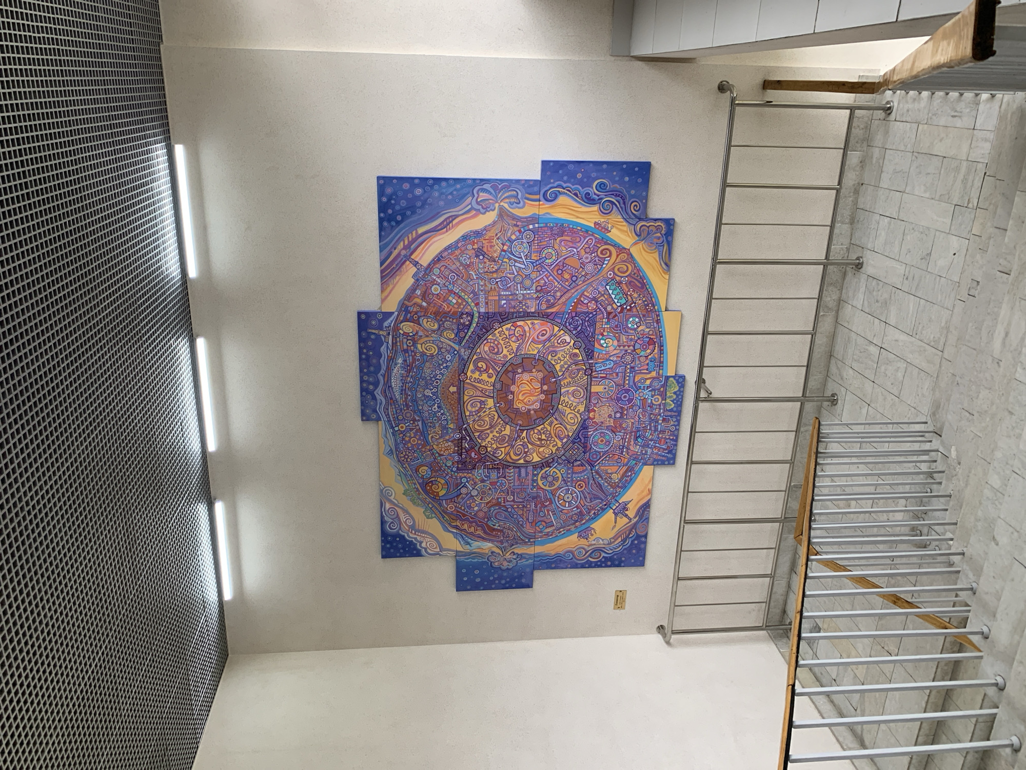 The image depicts an interior space. A large, colorful, mandala-style painting, composed of several canvases, is mounted on a white wall. To the left, a section of the wall is covered with a metal grid. Vertical lighting fixtures are present between the white wall and the metal grid. To the right of the painting, a metal ladder leans against the wall, next to a staircase with silver handrails and light-colored stone tiles on the wall. The floor appears to be white. The location is Moscow, Russia.