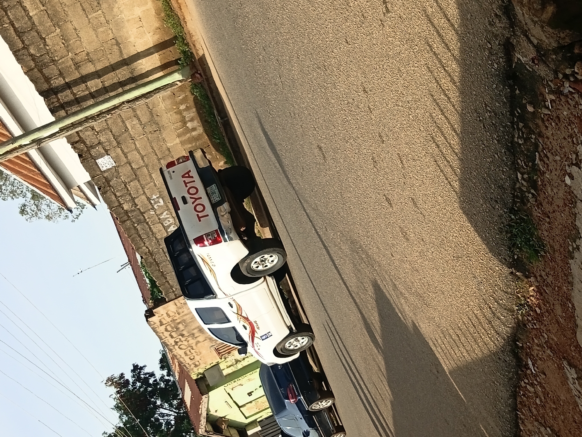 A white Toyota Hilux pickup truck and a dark blue sedan are parked on the side of a paved road. The pickup truck has "TOYOTA" written on the tailgate and a license plate that reads "FST 391 HY". It also features decorative decals on its side. The sedan is parked behind the pickup truck. The scene is framed by a brick wall and some buildings with corrugated metal roofs. The road is lined with sparse vegetation and debris. The sun casts long shadows across the road and wall, indicating it is likely late afternoon. The sky is clear and light blue.