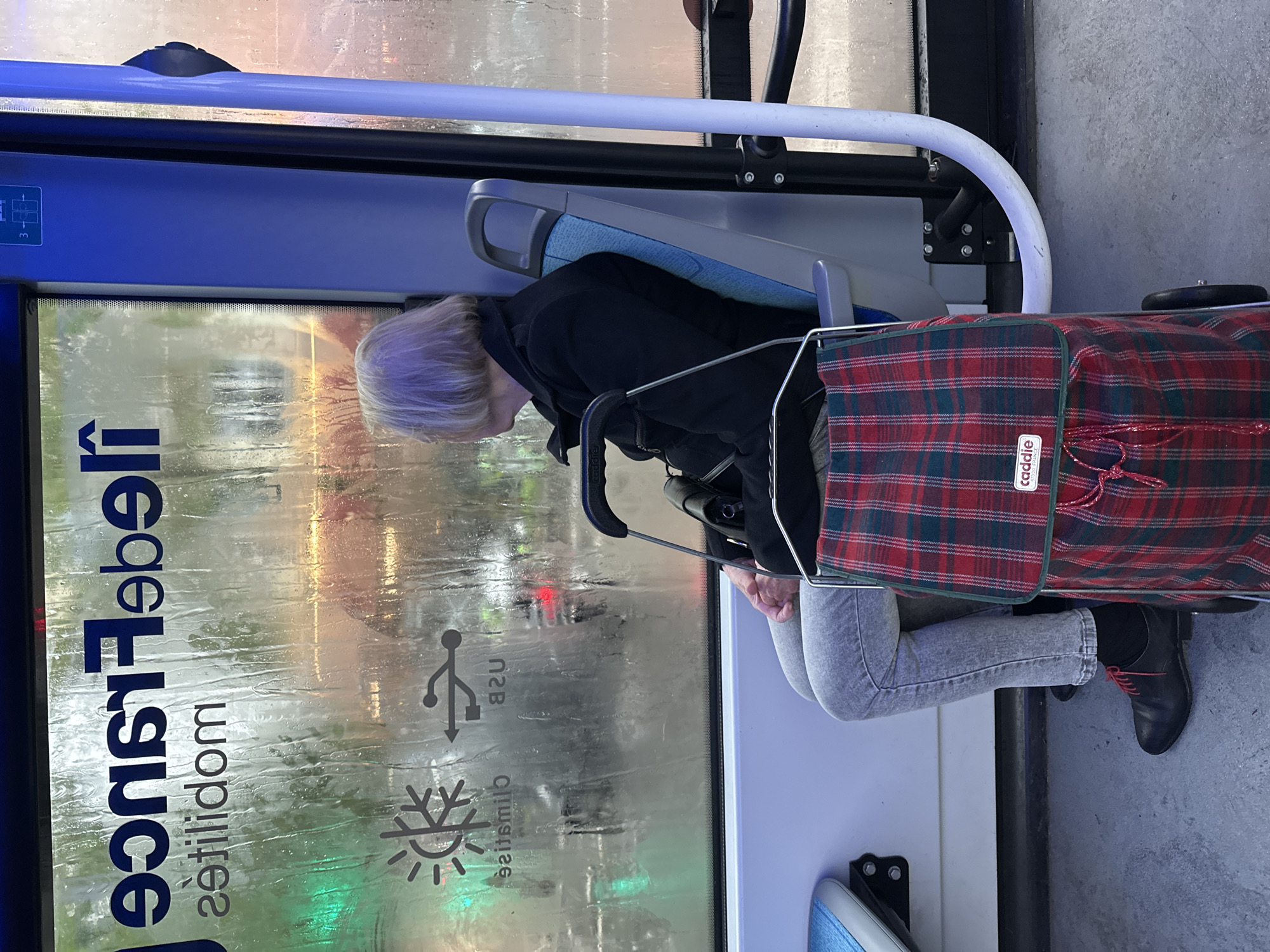A person sits on a bus in Paris, France. The bus window displays the words "Île de France Mobilités." The window is wet. The person is elderly and has light hair. They are wearing jeans, black shoes, and a black jacket. They have a red plaid bag on a rolling frame beside them. The bus is also marked with a USB charging station icon and an air conditioning icon.