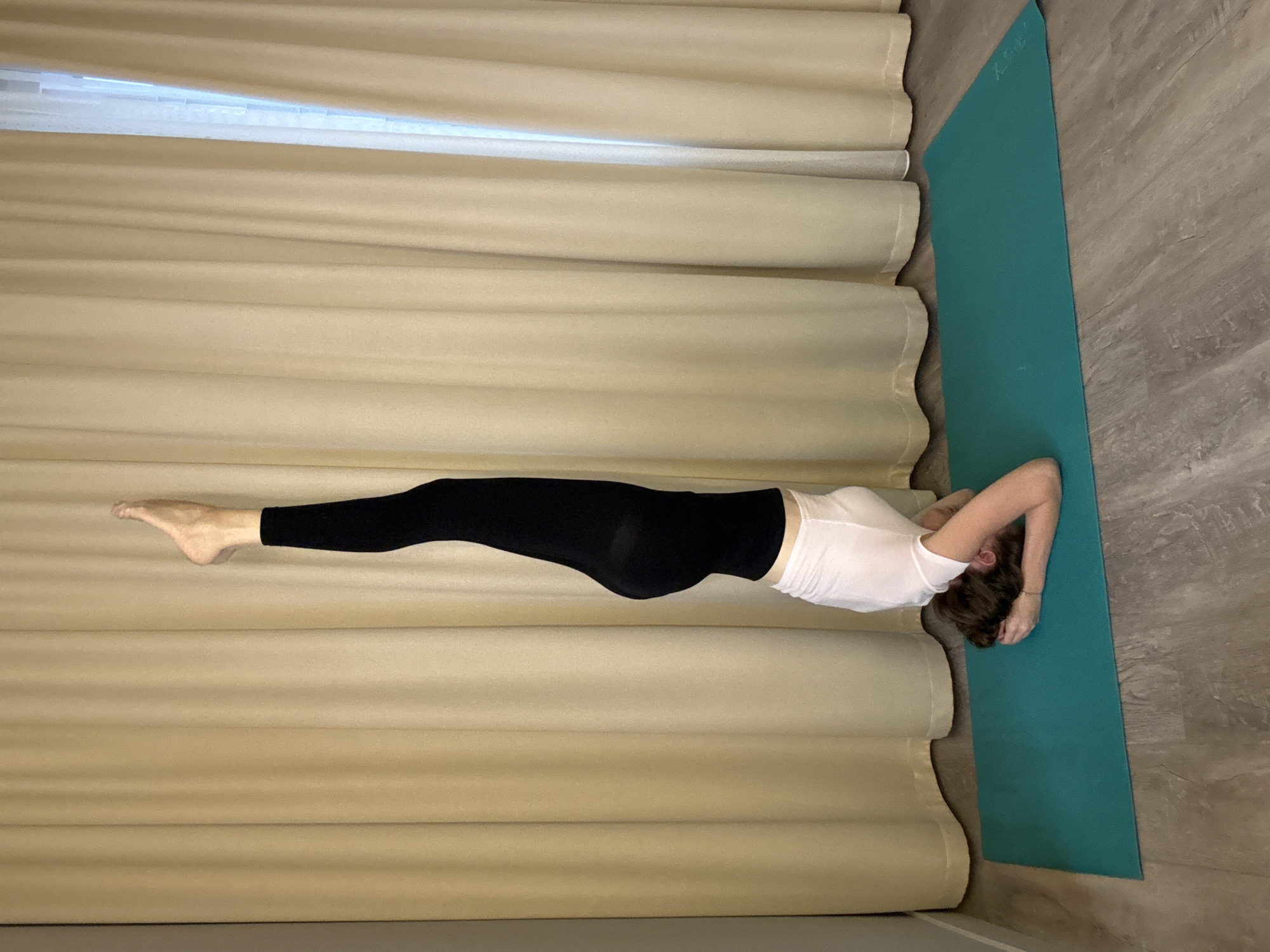 A person is performing a headstand yoga pose on a teal yoga mat. The person is wearing black leggings and a white t-shirt. Their head is on the mat, supported by their hands, and their legs are extended straight up in the air. The background consists of light beige curtains and a wooden floor. The scene is indoors. The image does not contain any information related to Dolgoprudny, Russia.