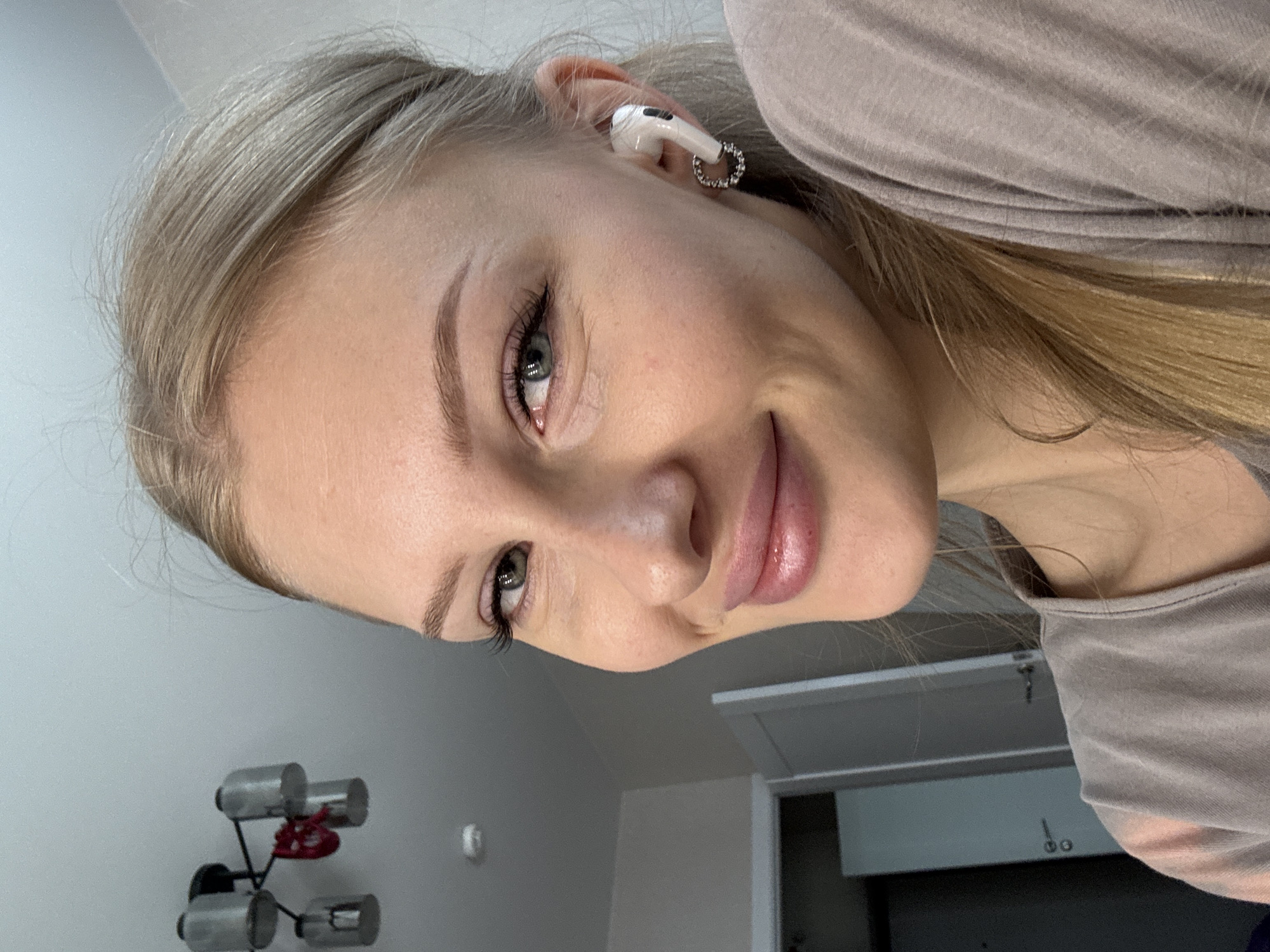 The image shows a close-up, selfie-style portrait of a young, fair-skinned woman with blonde hair tied back. She is wearing a light brown t-shirt and white wireless earbuds in her right ear. Her makeup includes eyeliner and lip gloss. The background is a lightly colored wall and a doorway. There is a light fixture with multiple bulbs visible on the ceiling in the upper left corner. The overall impression is of an indoor setting, possibly a home or office. The location of Perm, Russia, is not evident from the image itself.