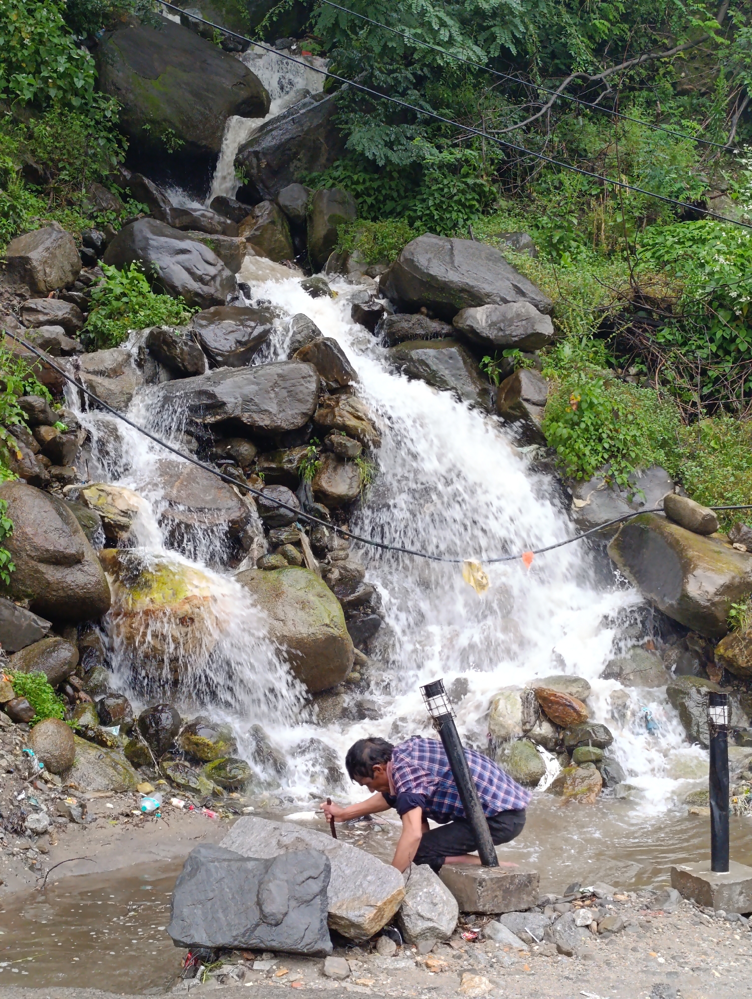 The image depicts a man in a plaid shirt and dark pants crouching by a waterfall in Vashisht, India. The waterfall flows over rocks, creating a natural cascade amidst lush green vegetation and large boulders. The man appears to be interacting with the water or the surrounding area. Two black lampposts are visible on the bank of the stream, suggesting a developed area near the natural feature. Debris, including plastic bottles and wrappers, is scattered on the ground near the water's edge, indicating a potential environmental concern. The overall scene captures a moment of human activity in a rugged, natural setting.