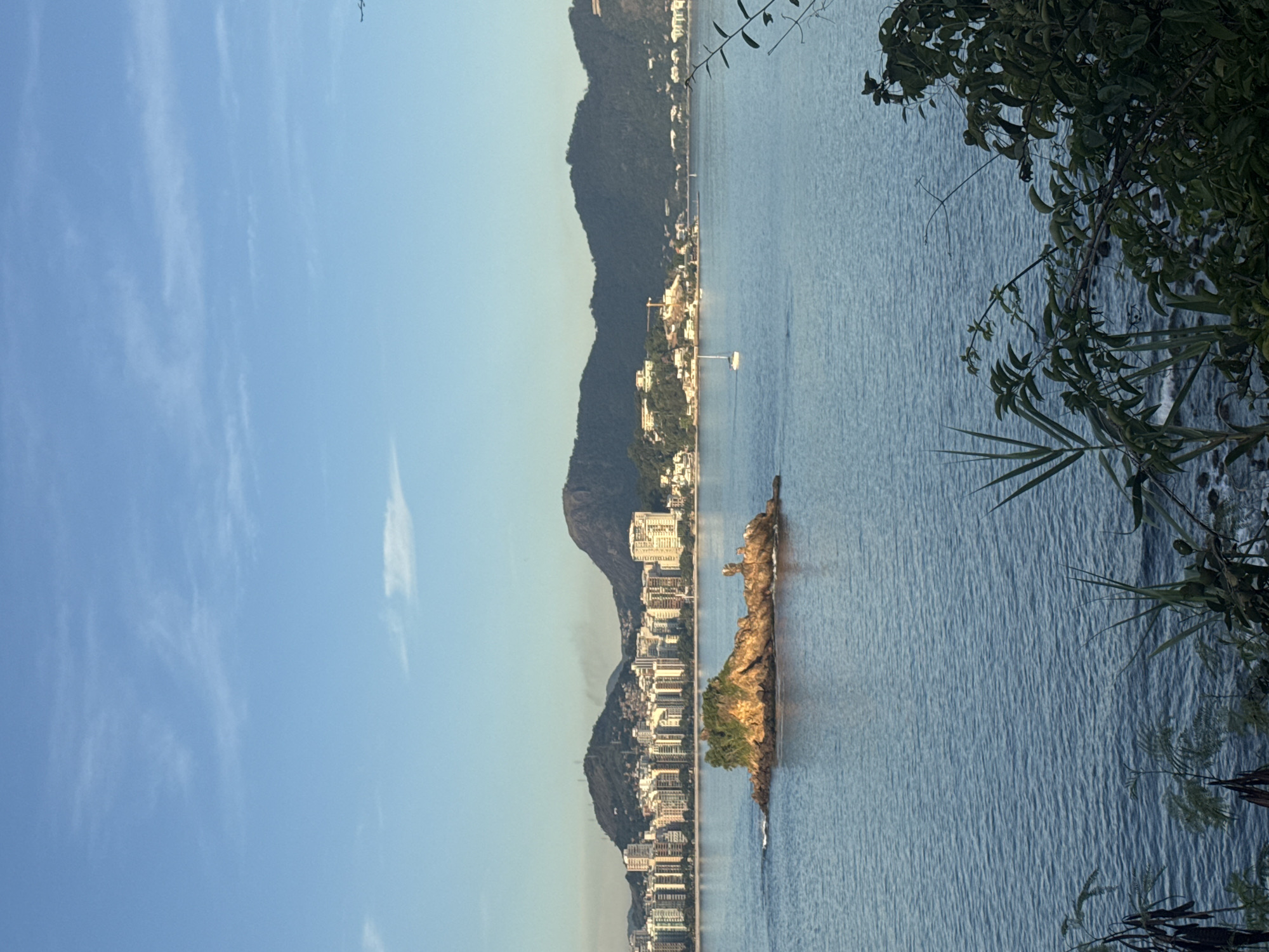 This is a photograph of the Icarai beach in Niterói, Brazil, taken at sunrise. The image captures a panoramic view of the Guanabara Bay, with a small, rocky island in the foreground. The island is covered with lush greenery and has a few small structures on it. In the background, the cityscape of Niterói unfolds, with its characteristic buildings and a backdrop of lush, green mountains. The sky is a clear blue with a few wispy clouds, and the sun is just beginning to rise, casting a warm glow over the scene. A single sailboat is visible on the water, adding a touch of serenity to the tranquil setting. The foreground features some foliage, suggesting the photo was taken from a vantage point overlooking the bay.