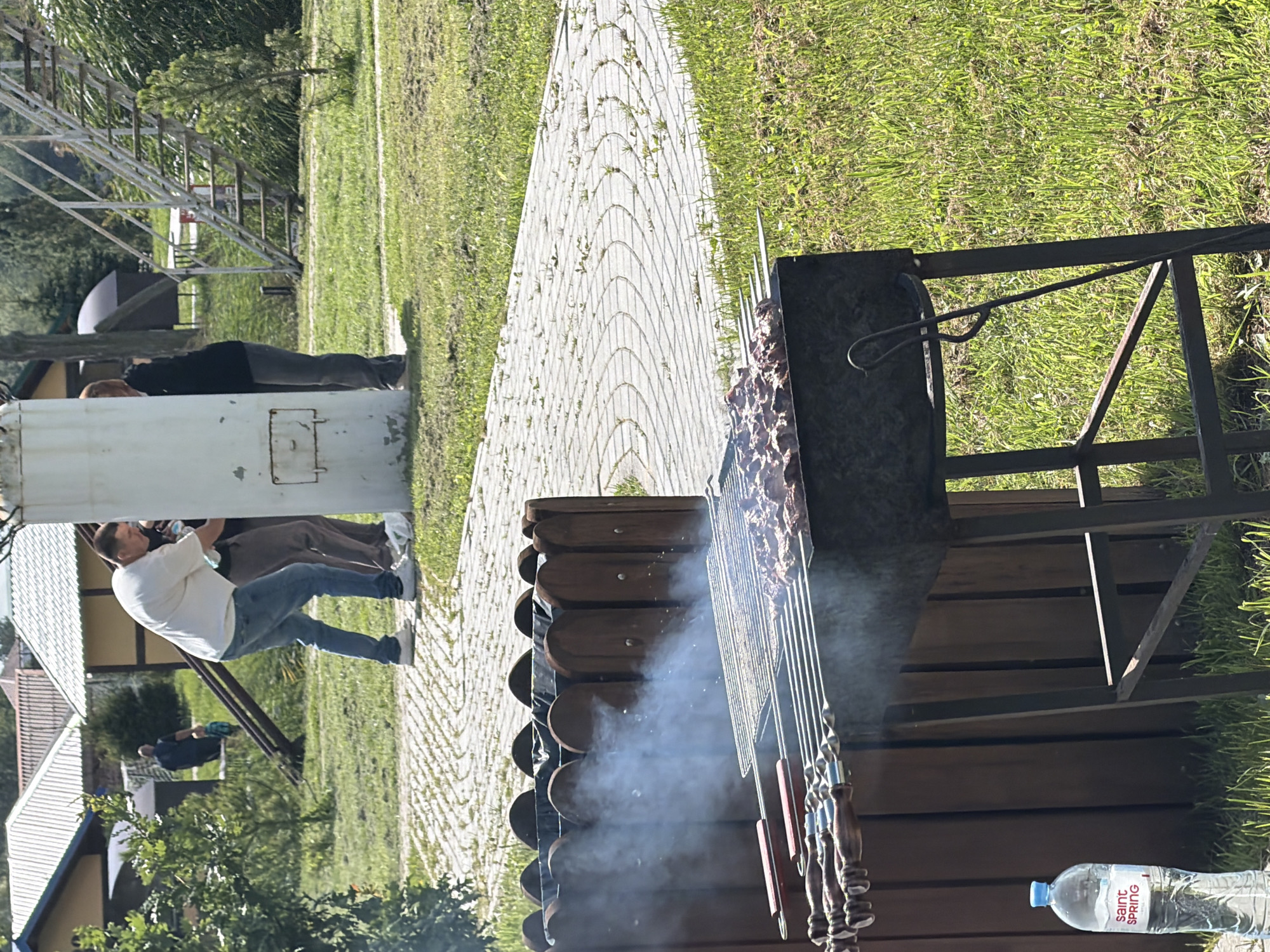 In an outdoor recreational area in Konstantinovka, Russia, three individuals are present. One man, wearing a white t-shirt and blue jeans, is leaning forward, his face obscured as he appears to be interacting with a large, white cylindrical object. Another person, in a black t-shirt and grey pants, stands beside him, partially obscured. A third individual, wearing dark clothing, is visible in the background near a set of metal stairs.
In the foreground, a metal barbecue grill is actively cooking skewers of meat, producing visible smoke. Beside the grill, a wooden trash receptacle is positioned. To the left of the grill, a plastic water bottle labeled "Saint SPRING" stands upright on the grass. The scene is set on a sunny day with a paved pathway in the mid-ground and a grassy area surrounding the barbecue. Buildings with pitched roofs are visible in the background.