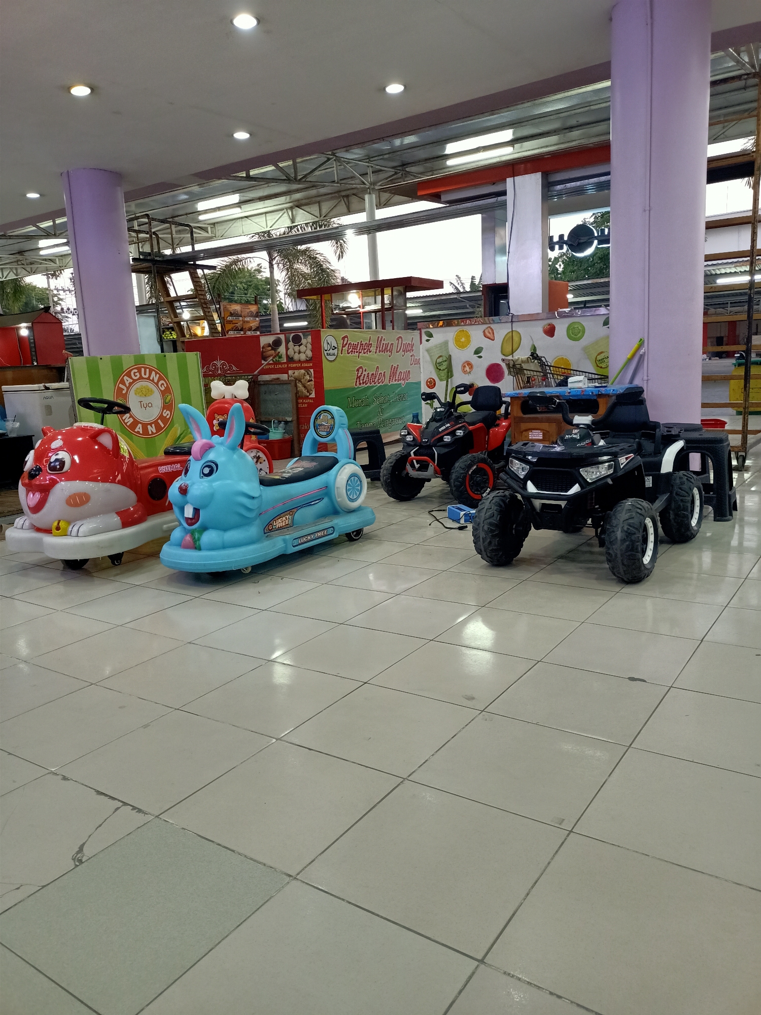 The image depicts a shopping area in Mernung, Indonesia, featuring a row of children's ride-on toys and food stalls. In the foreground, two brightly colored ride-on toys are prominently displayed: a red fox-shaped vehicle and a blue bunny-shaped vehicle. Behind these, two quad bike-style ride-on toys, one red and one black, are parked. The background reveals food stalls with banners advertising various food items, including "Jagung Manis" (sweet corn) and "Pempek Lingah Dyah." The flooring is tiled, and overhead lighting illuminates the scene. The overall impression is that of a vibrant, family-friendly commercial space.