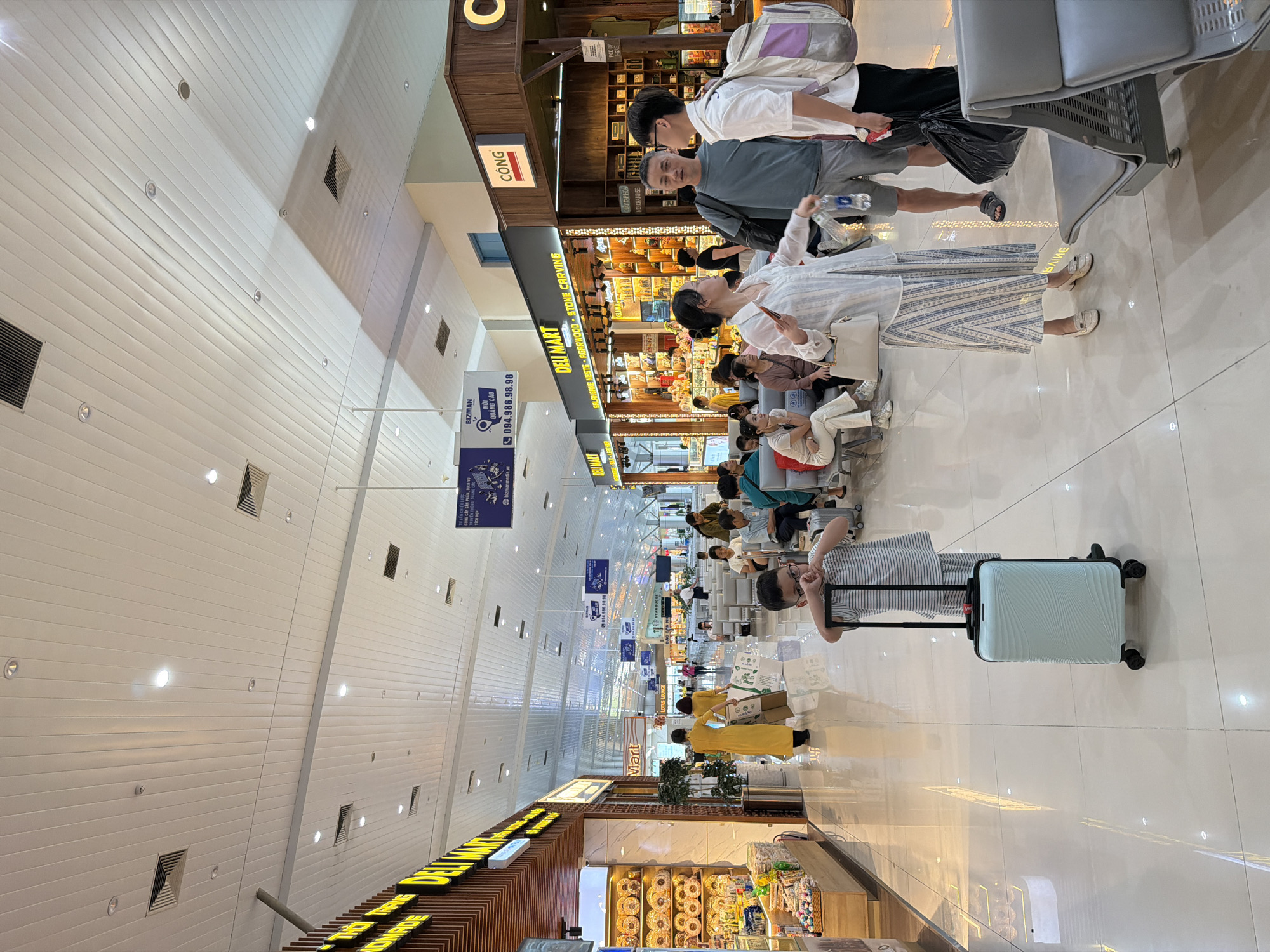 The image depicts the interior of an airport terminal, specifically within the departure lounge area of Cam Ranh International Airport in Cam Ranh City, Vietnam. The scene is brightly lit with fluorescent lighting from the long, white, paneled ceiling. Various shops, including a "DELI MART" and another labeled "CÔNG," are visible along the right side of the terminal. People are seen sitting on grey airport seating benches, waiting.
In the foreground, a young boy wearing a striped t-shirt and shorts stands next to a light blue rolling suitcase. To his right, a woman in a white and blue striped dress, holding a white handbag and a bottle of water, is engaged in conversation with two men. One man, wearing a grey t-shirt and shorts, carries a black bag. The other man, in a white shirt and shorts, has a backpack slung over his shoulder. Further back, a woman in a yellow dress and another in a similar yellow outfit with shopping bags are walking away from the camera. The floor is a polished, light-colored tile. The overall atmosphere suggests a typical, busy airport environment.