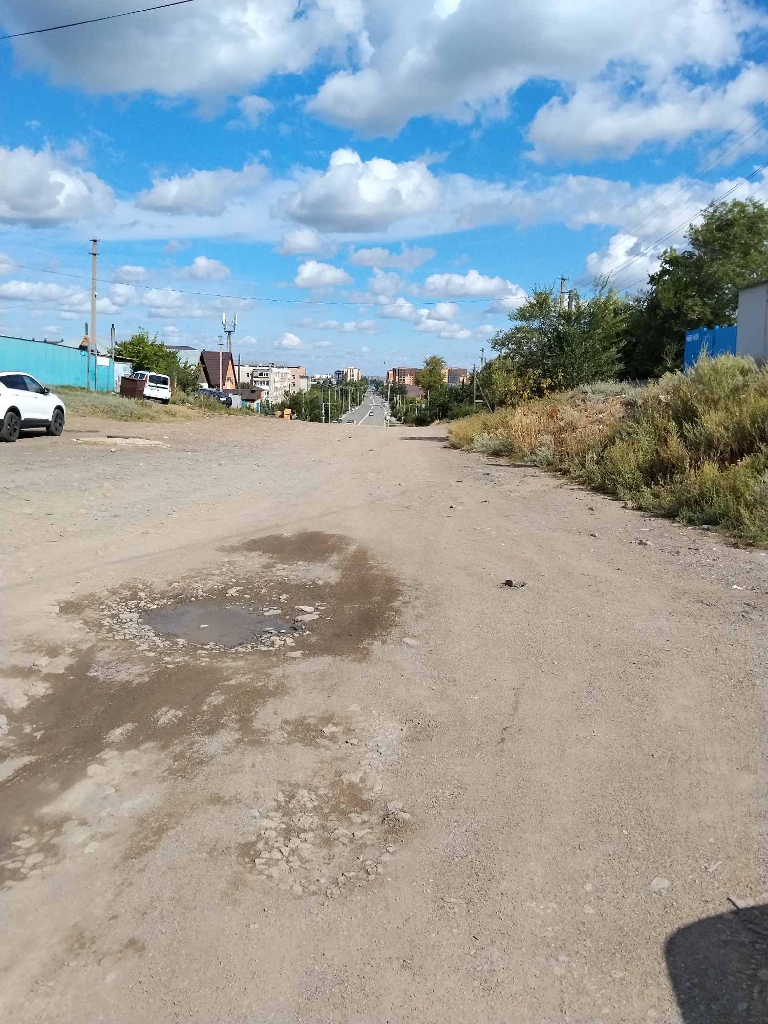This is an outdoor, ground-level photograph taken on a dirt road in Kokshetau, Kazakhstan. The road is unpaved and appears to be in poor condition with several large puddles and potholes. The road slopes downwards into the distance where it appears to transition into a paved road lined with buildings and trees. The sky is blue with scattered white clouds. A white car is parked on the left side of the road, and several other vehicles are visible further down the road. There are also some buildings and fences visible in the background. The overall scene depicts a somewhat neglected urban or semi-urban environment.