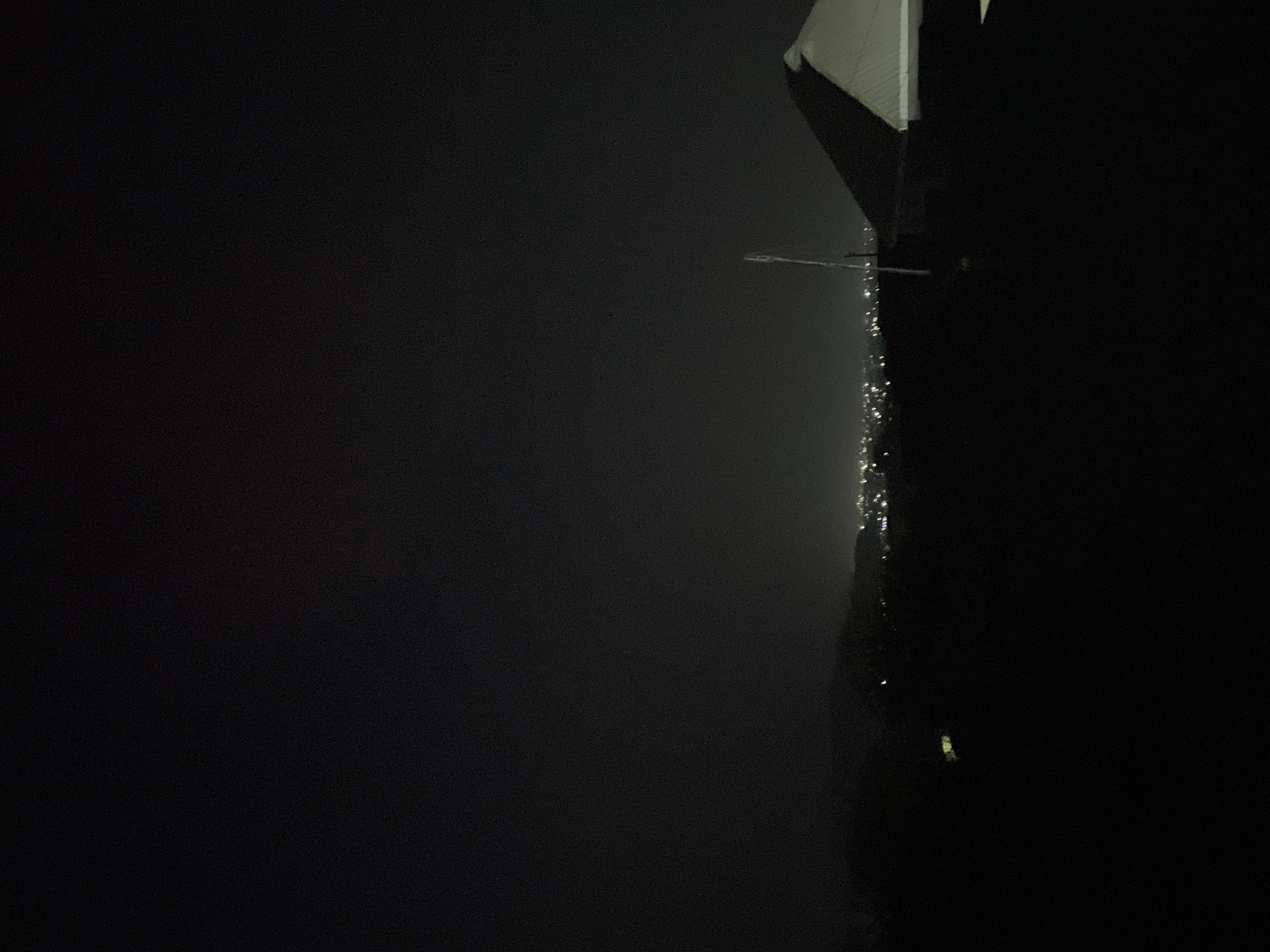 A low-angle, nighttime, long-exposure photograph shows a dark scene. The upper-right corner features the edge of a white roof. Directly to the left of the roof, running vertically, is a series of small, blurry lights that likely represent city lights in Jos, Nigeria. The majority of the scene is obscured by darkness, suggesting fog or a lack of light. A streetlight appears as a blurry object. The absence of distinct details and the darkness obscures most contextual information.
