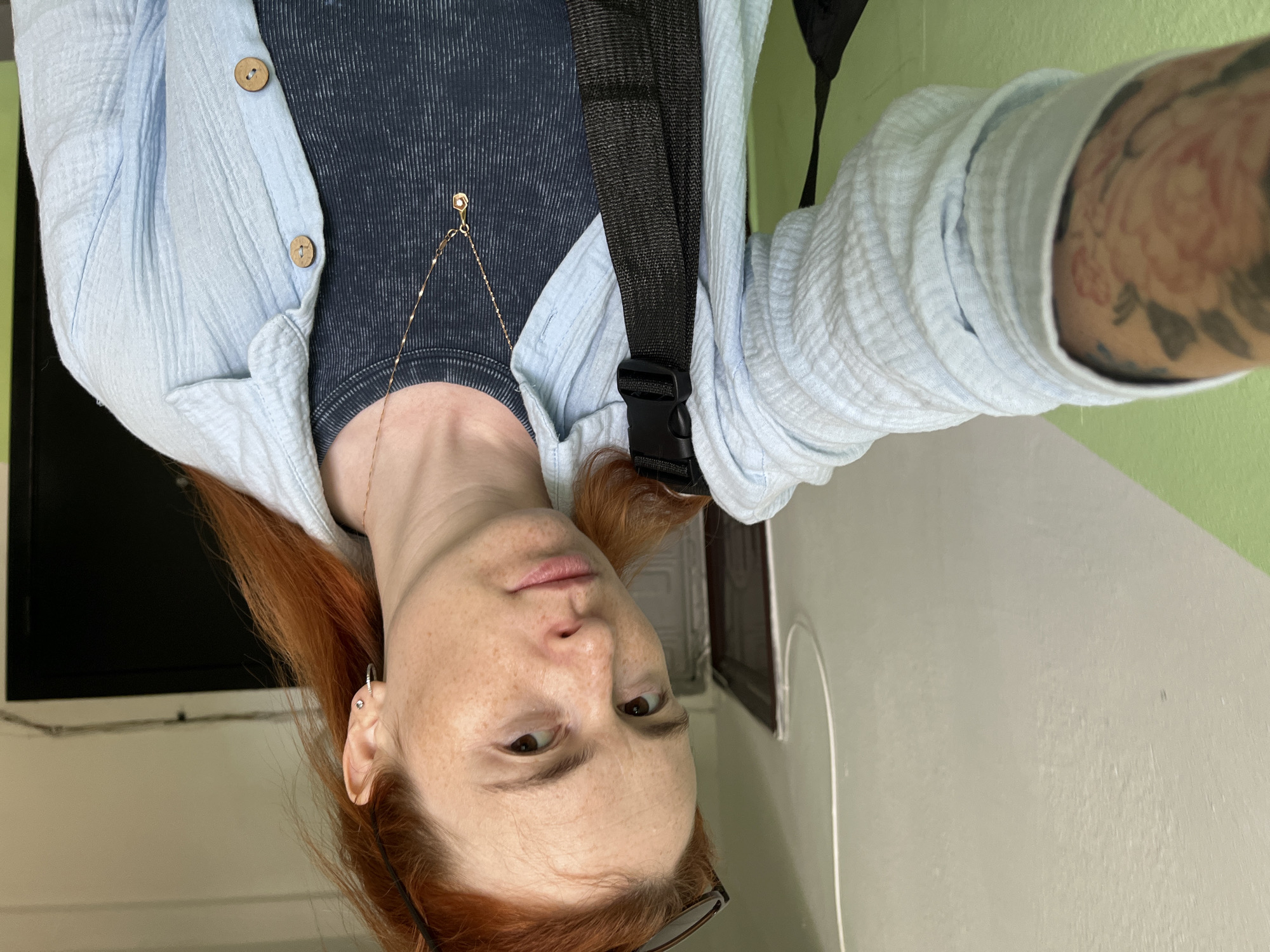 A young woman with fair skin, freckles, and red hair is taking a selfie. She is wearing a light blue, textured shirt over a dark blue ribbed t-shirt and a gold necklace with a hexagonal pendant. She has a black strap from a bag across her chest. Her left arm, visible on the left side of the frame, has a large floral tattoo. She is standing in a hallway with pale green and cream-colored walls. A white cord runs along the wall near the ceiling, and a black door is visible in the background on the right. This appears to be a residential hallway, possibly in Chaikovsky, Russia, given the information provided about the location.
